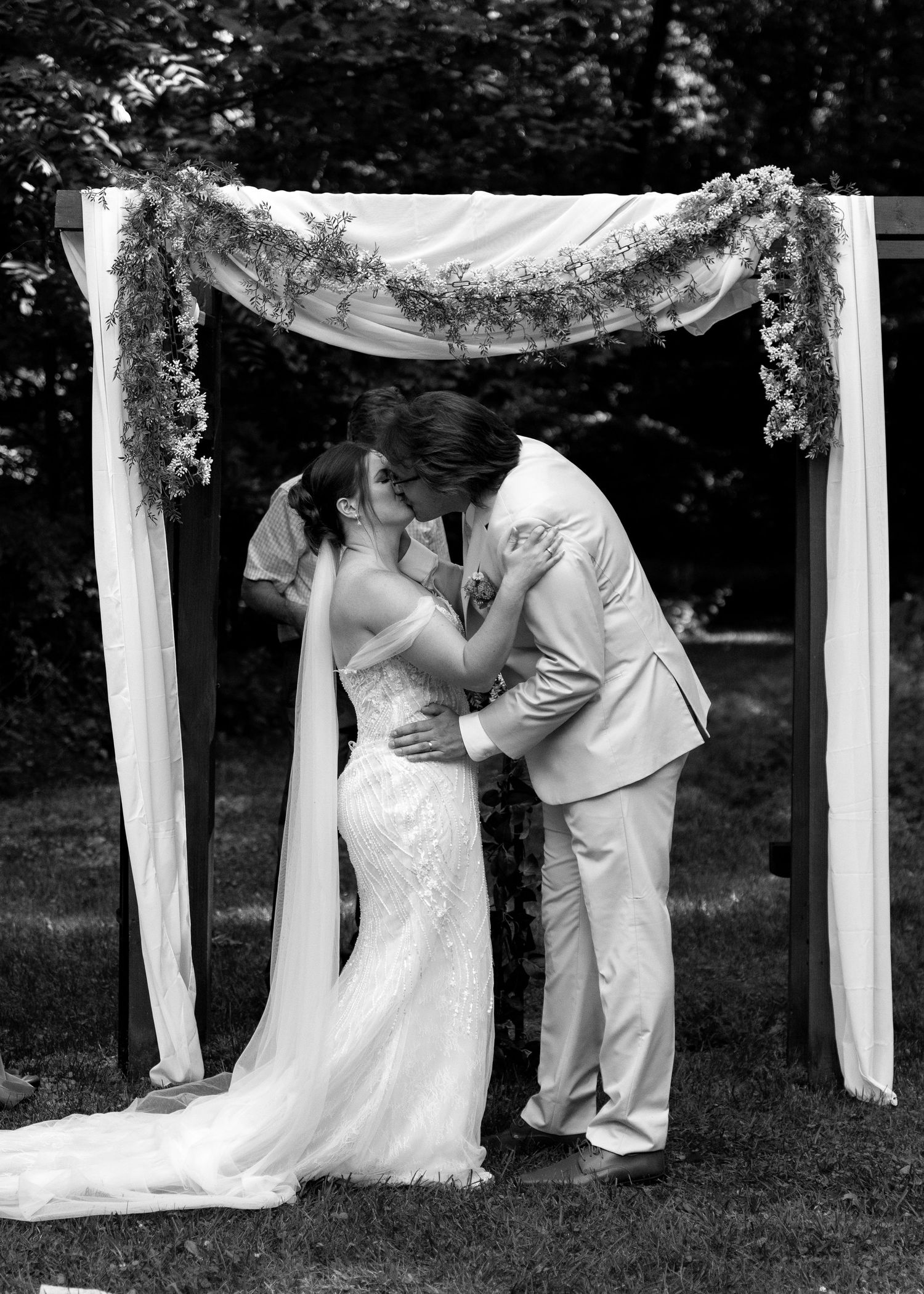 Black and white photo of a bride and groom sharing their first kiss under a floral arch at an outdoor wedding ceremony.