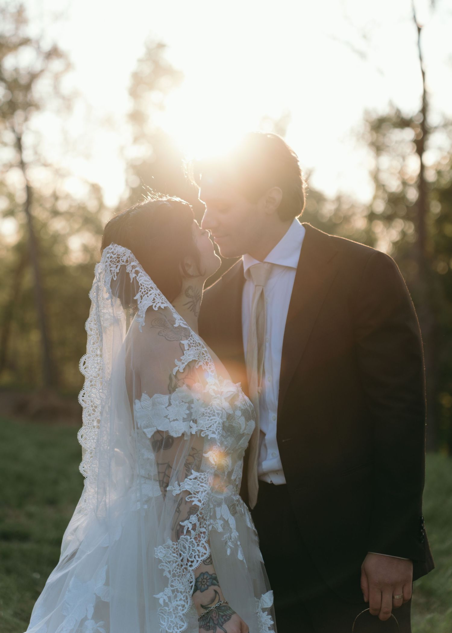 Bride and groom share a golden hour kiss, backlit by warm sunlight, bride wearing floral lace veil.