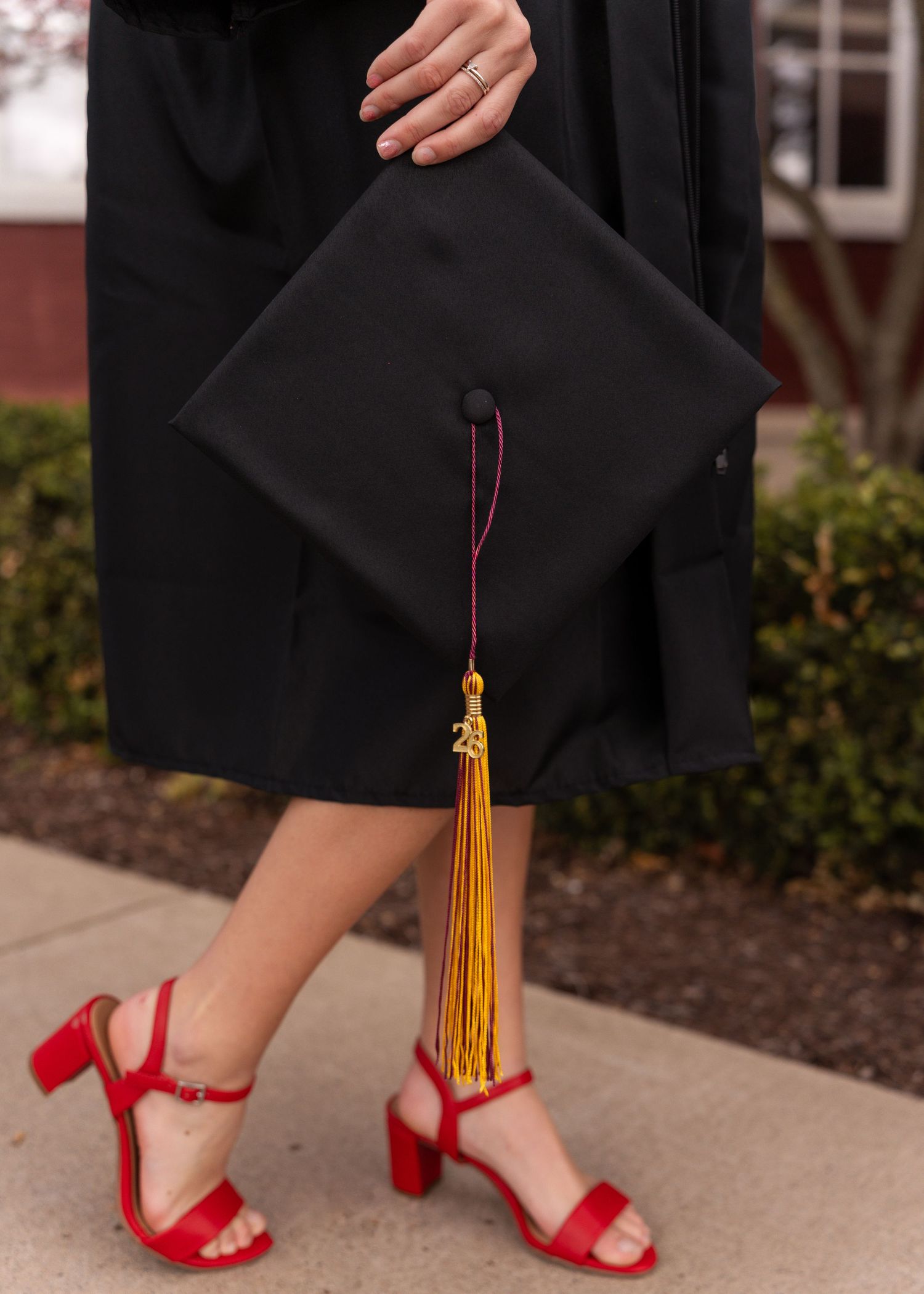 Graduate in black cap and gown holding mortarboard with gold tassel wearing red heels.
