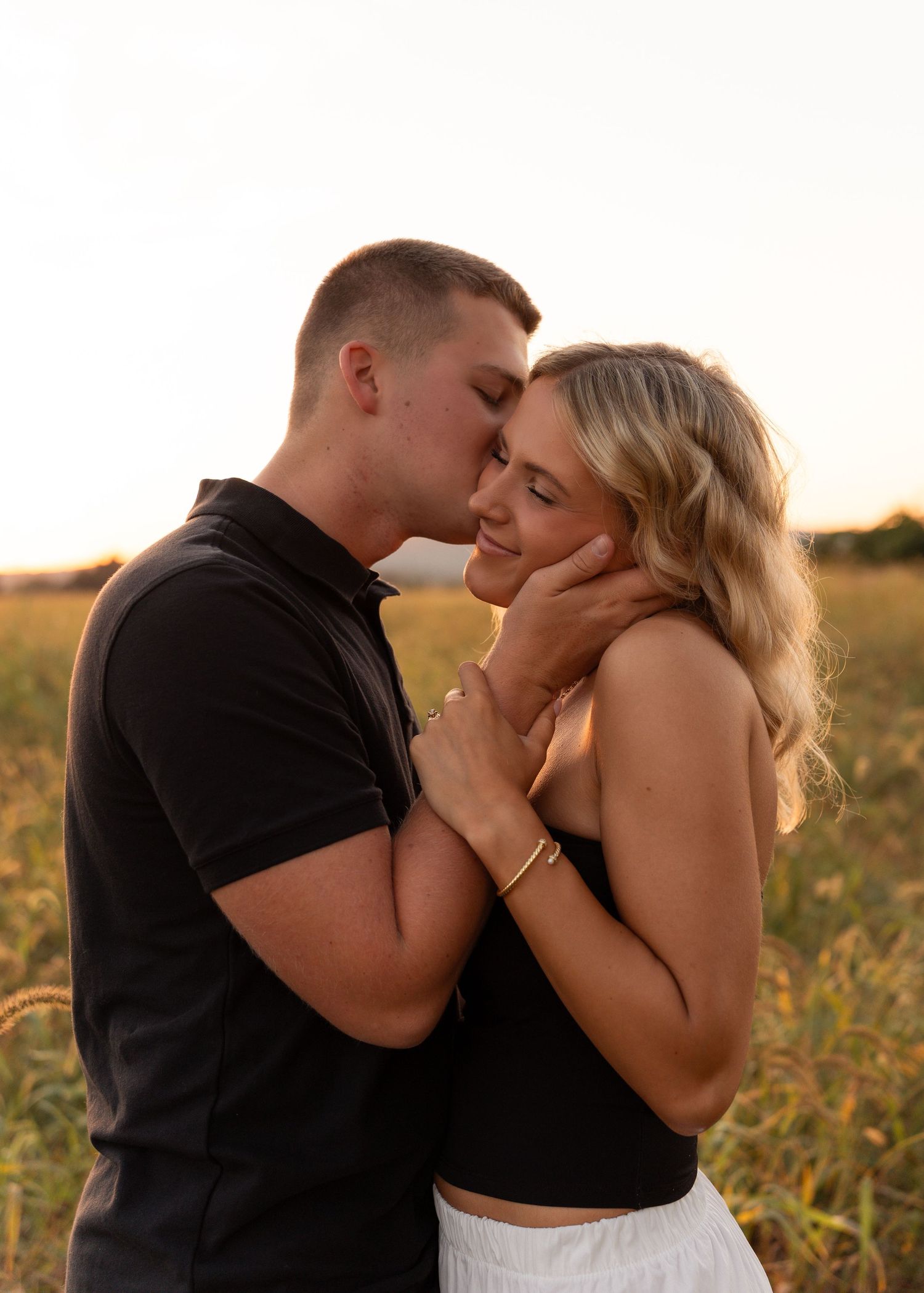A couple shares a romantic kiss in a sun-drenched field at golden hour, man gently cradling woman's face.