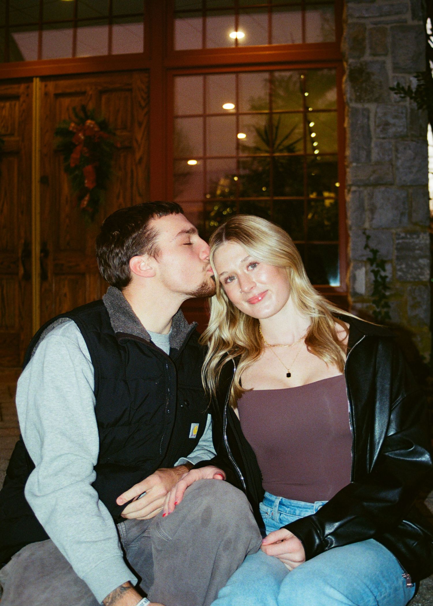 Couple sitting together outdoors at night, man kissing woman on cheek near decorated entrance.