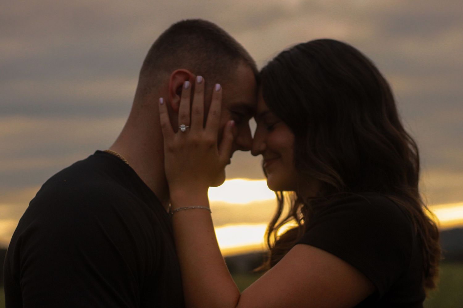 Engaged couple touches foreheads tenderly at sunset, woman displaying diamond engagement ring on hand.