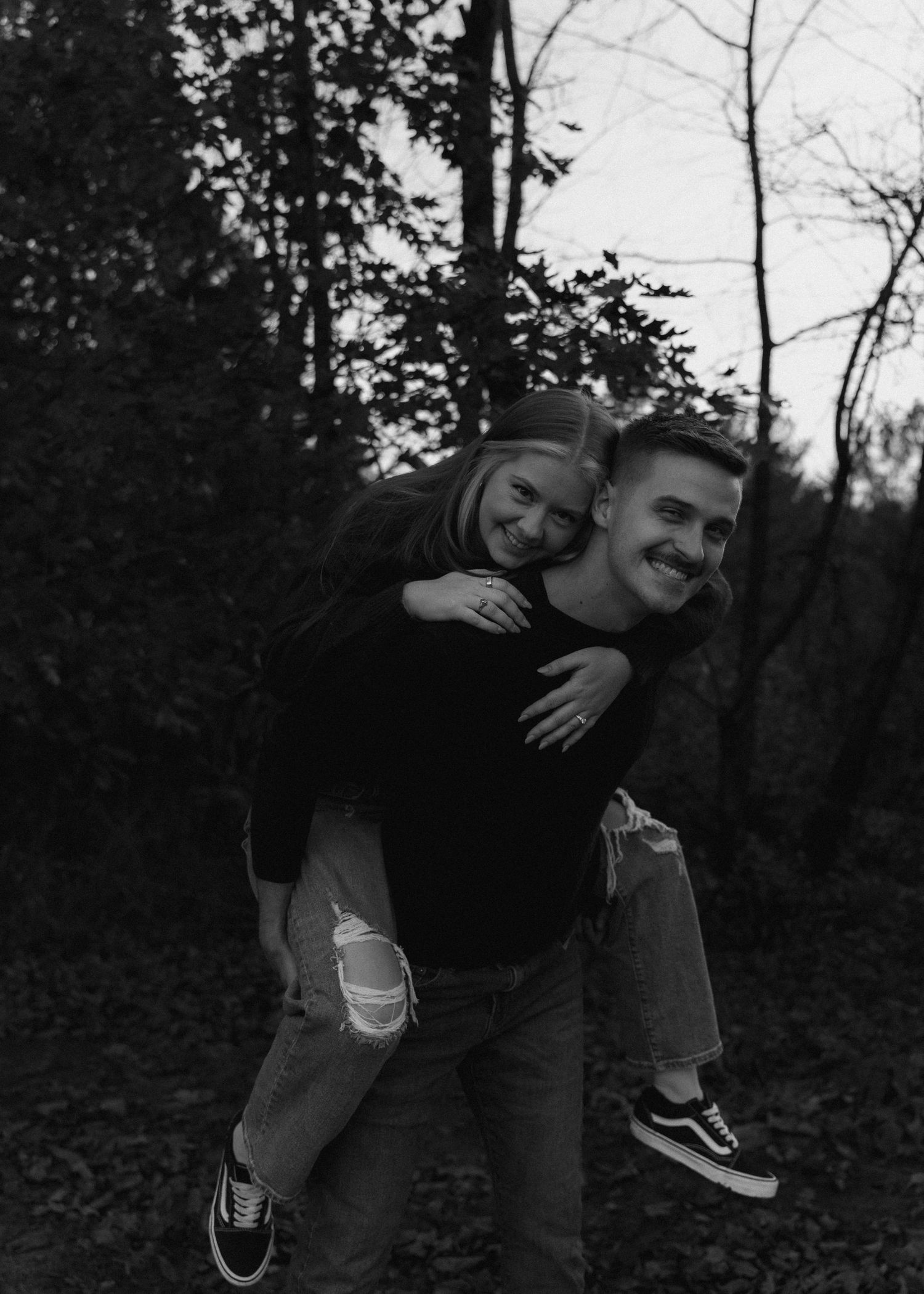 Black and white photo of a couple laughing as man gives woman a piggyback ride in an autumn forest.