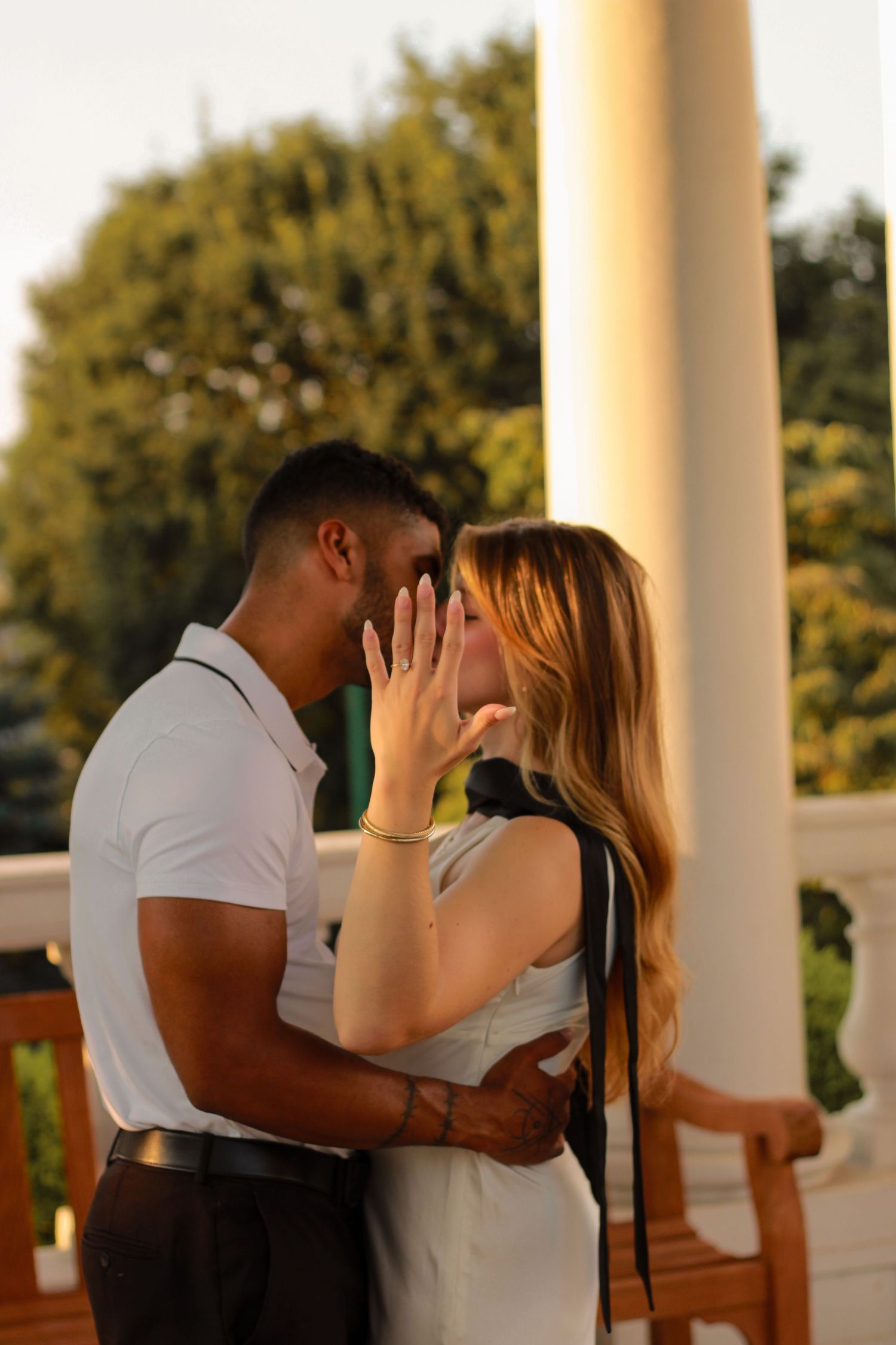 Couple sharing a kiss at golden hour on a porch, woman showing her engagement ring.