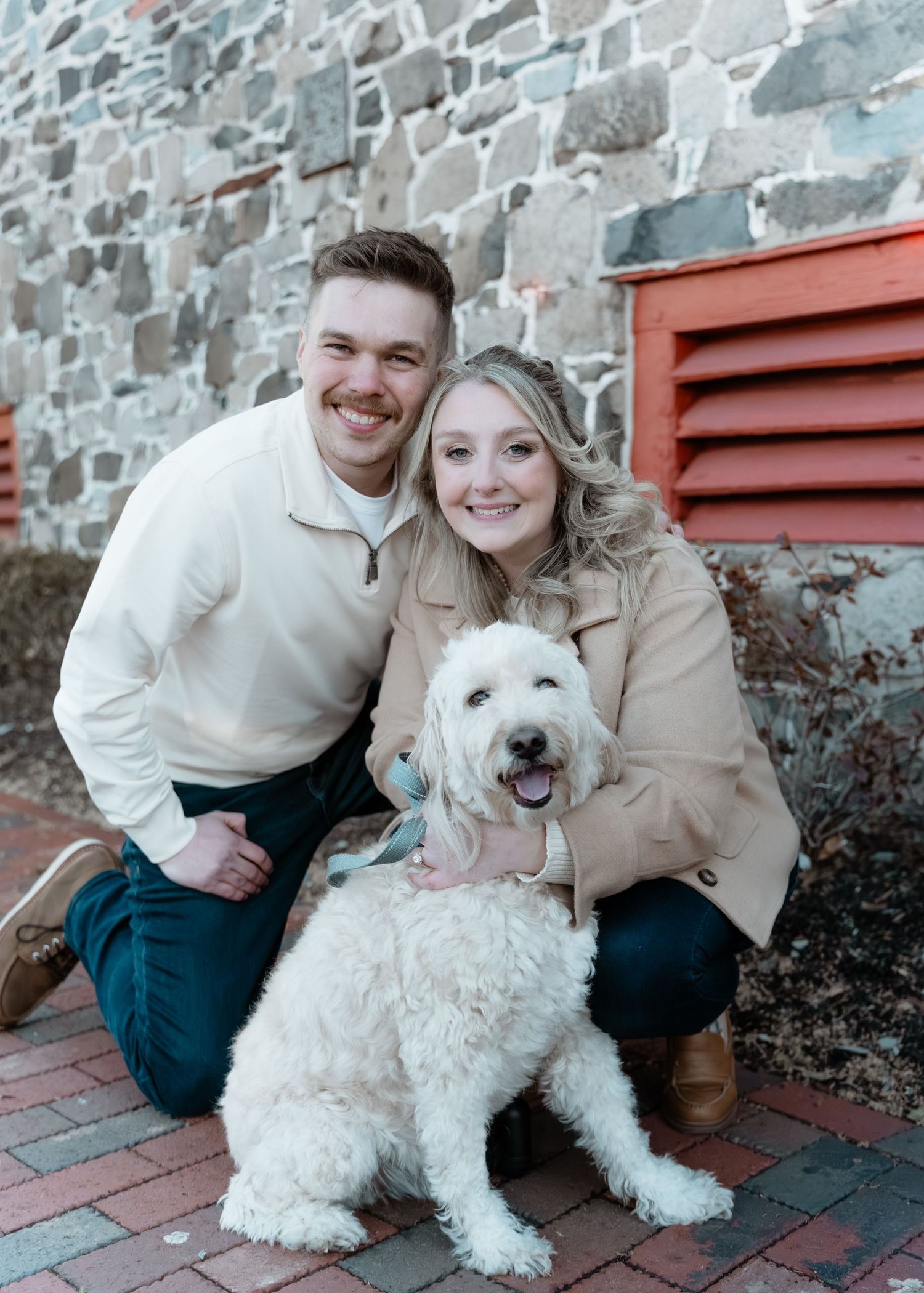 Smiling couple posing with their fluffy white dog in front of a rustic stone wall outdoors.