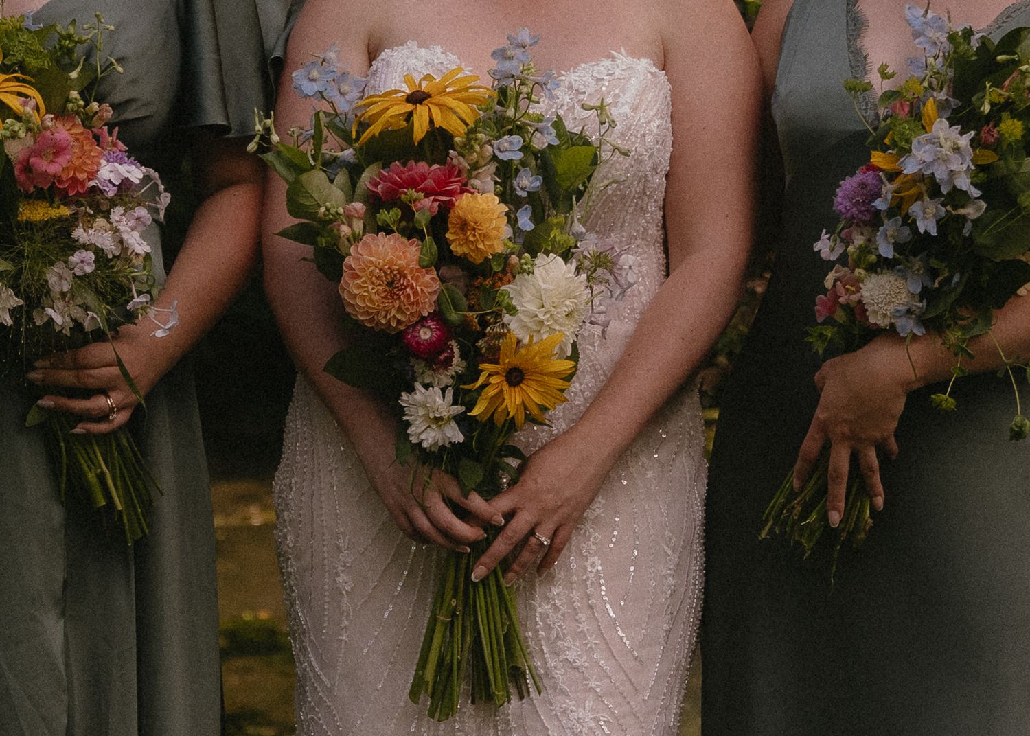 Bride in beaded gown holding vibrant sunflower bouquet flanked by bridesmaids in sage green dresses.