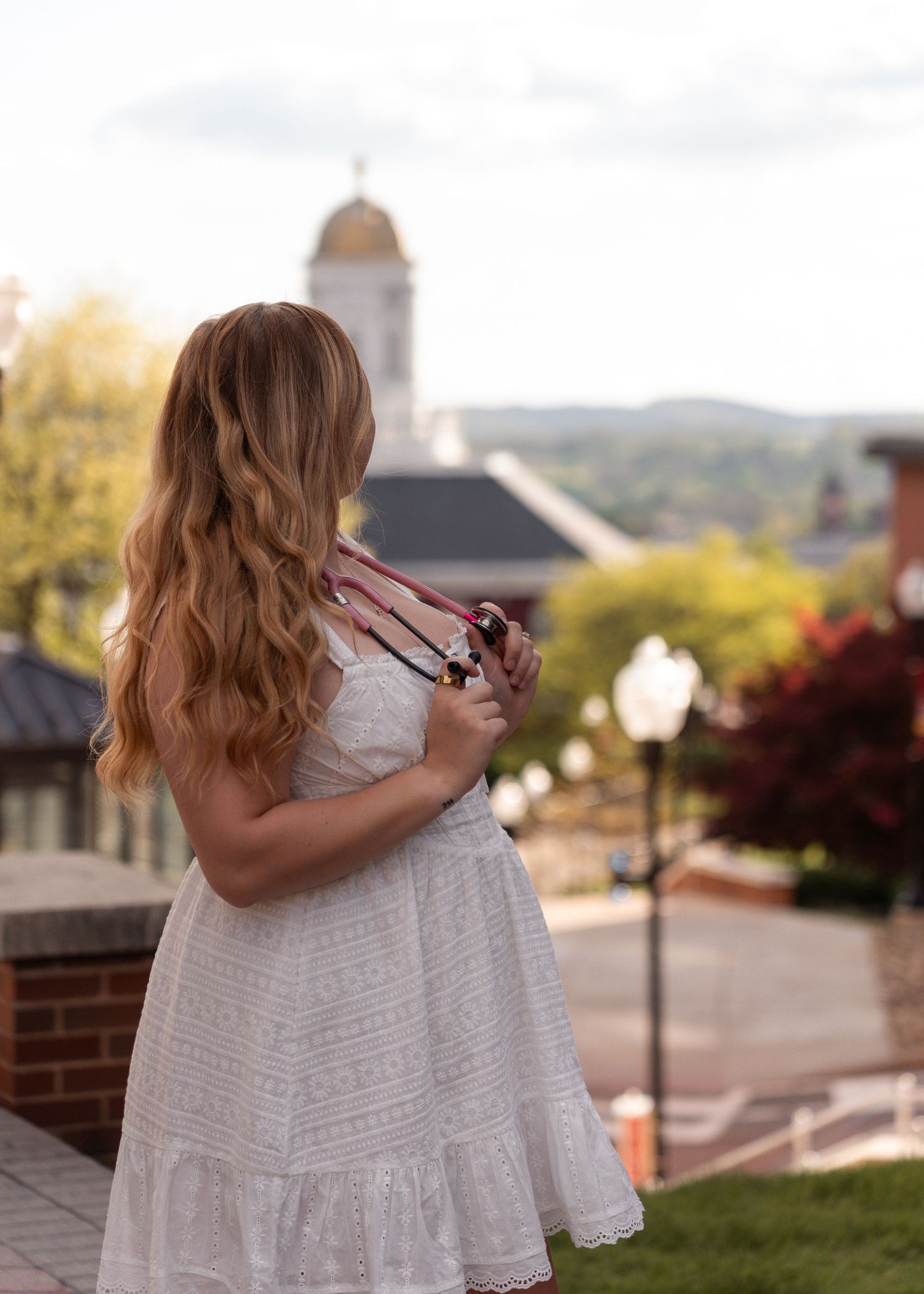 Young woman in white dress holding graduation regalia, standing with her back to camera near a domed building.