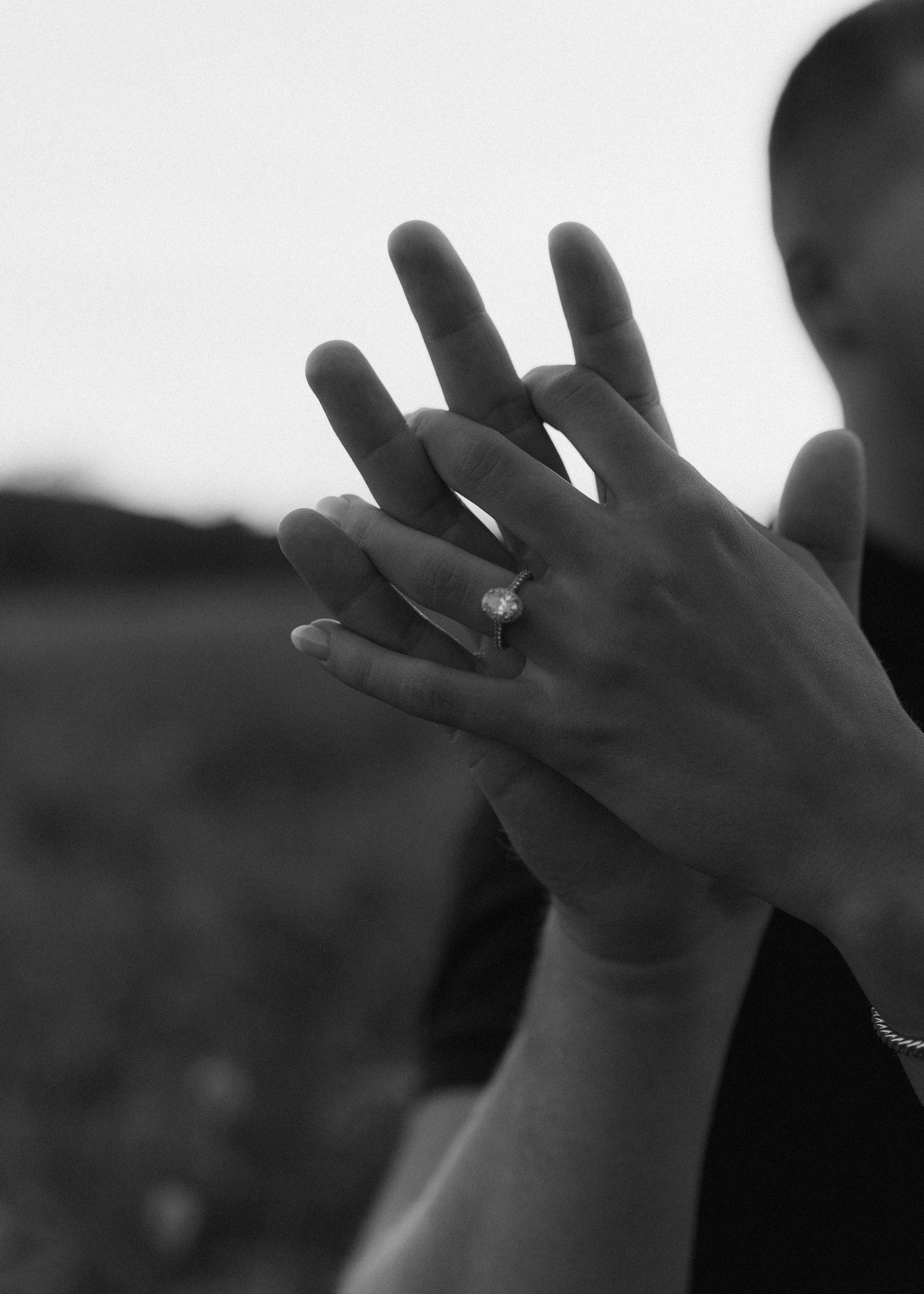 Black and white close-up of two hands intertwined, highlighting a sparkling diamond engagement ring.