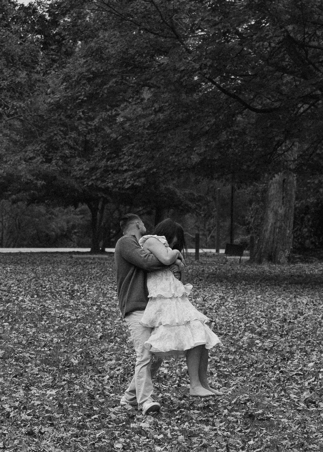Black and white photo of a couple dancing and embracing in a leaf-covered park in autumn.
