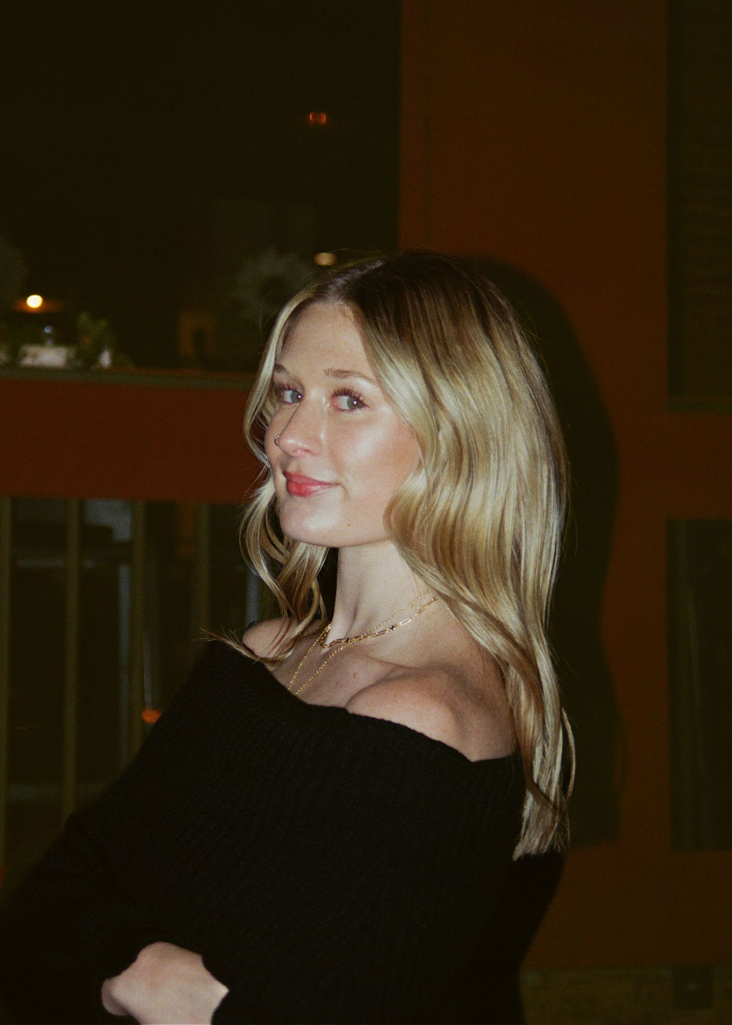 Blonde woman in black off-shoulder top posing indoors with warm ambient lighting.