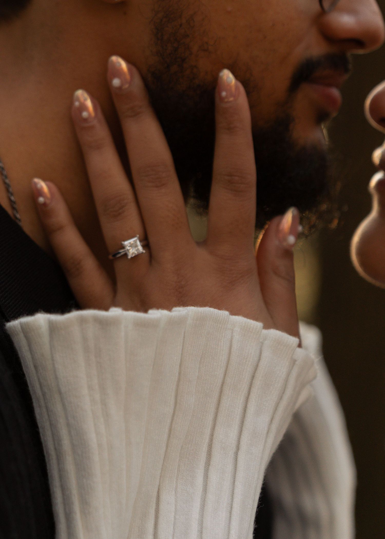 Close-up of womans hand showing square diamond engagement ring resting on mans bearded face.