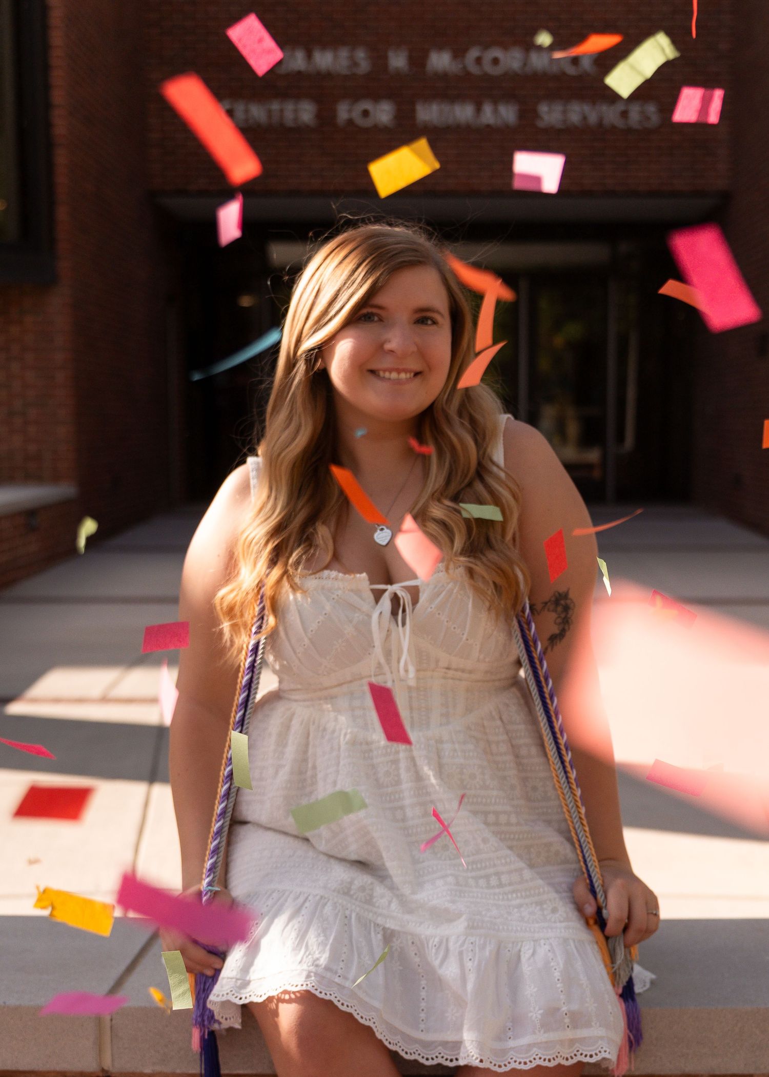 Smiling graduate in white dress with colorful confetti flying outside James H. McCormick Center for Human Services.
