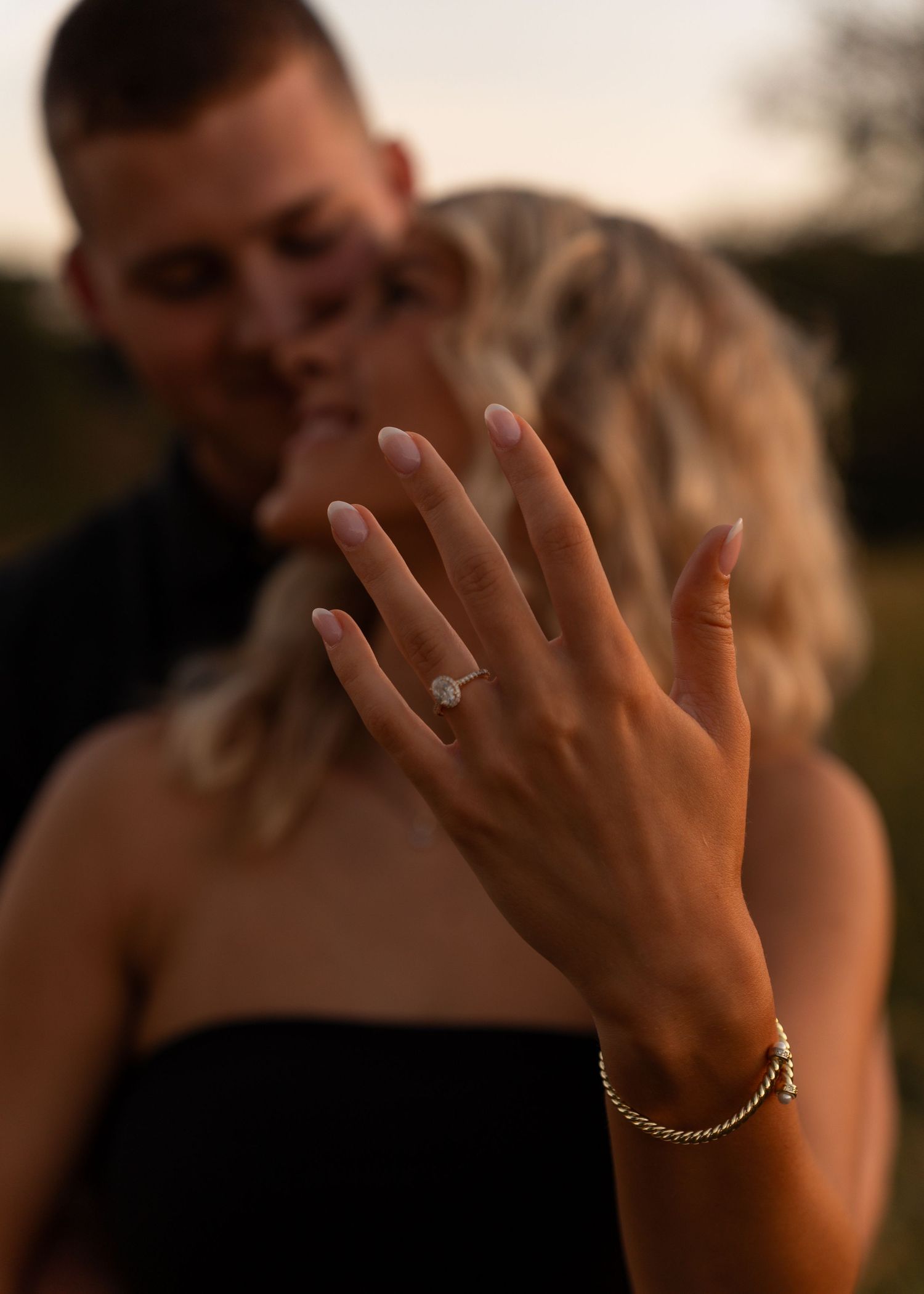 Engaged woman displays her elegant halo diamond ring in focus while couple kisses softly at golden hour.
