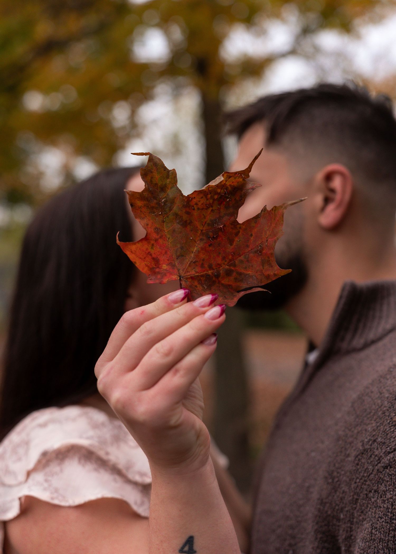 Couple sharing a kiss behind a vibrant red autumn maple leaf held up by the woman.