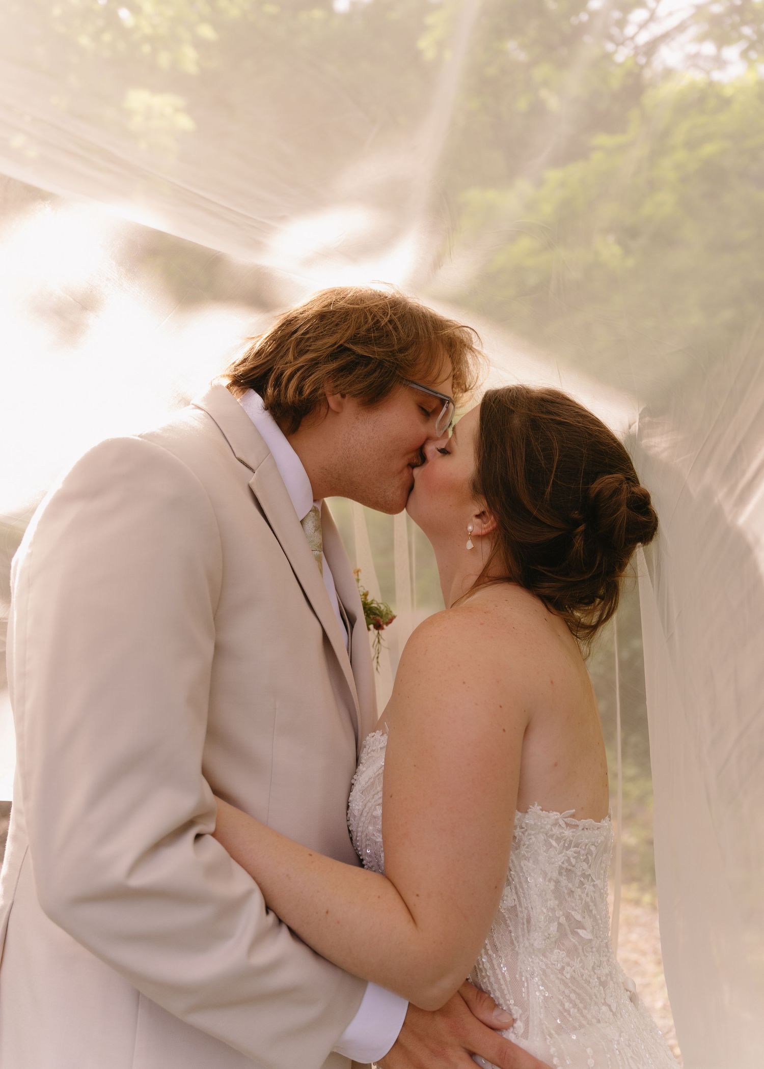 Bride and groom sharing a romantic kiss under a flowing veil outdoors on their wedding day.