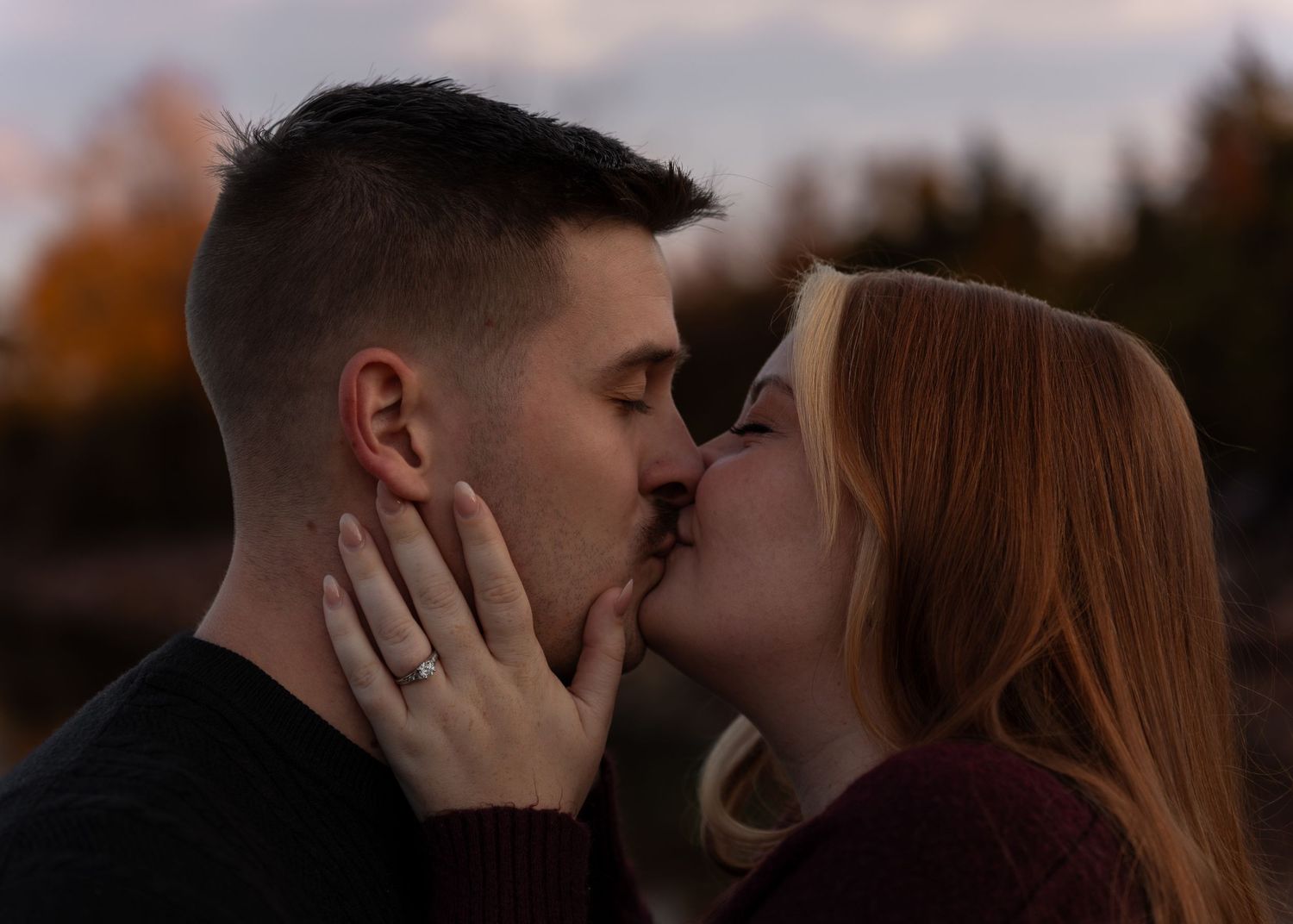 Couple sharing a romantic kiss outdoors during autumn sunset with colorful foliage behind them.