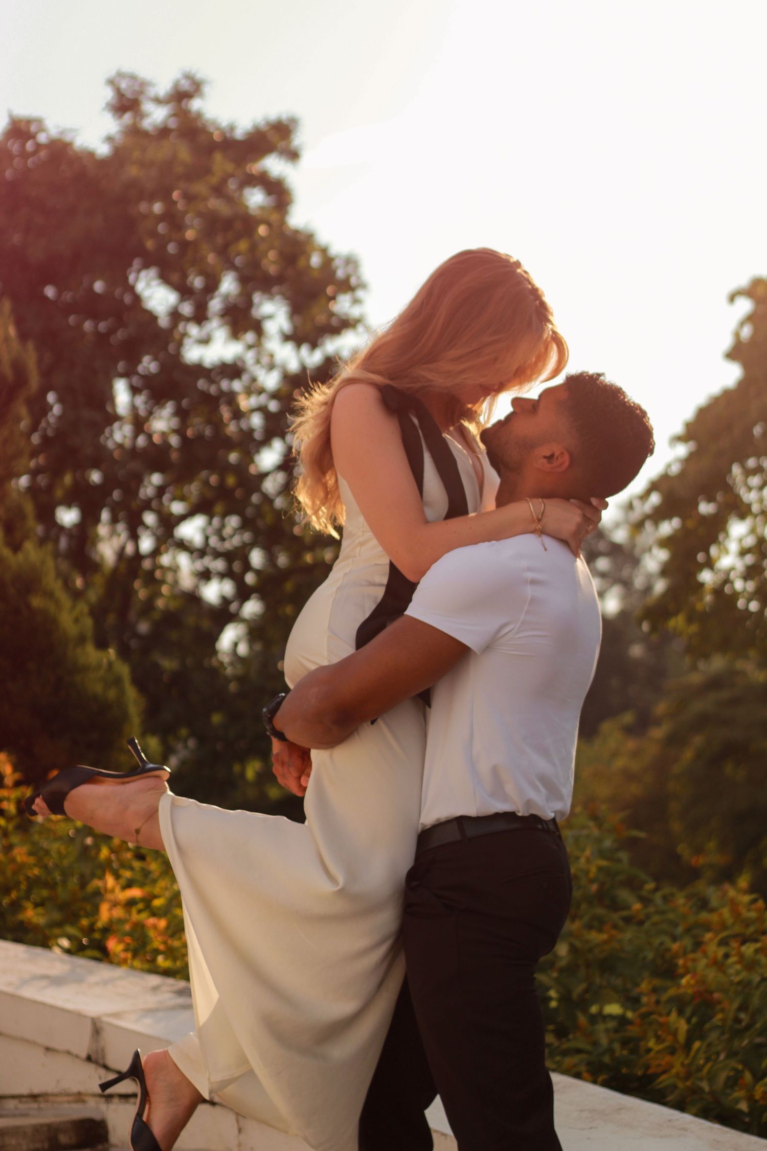 Romantic couple kissing at sunset in a lush garden, man lifting woman in a flowing white dress.