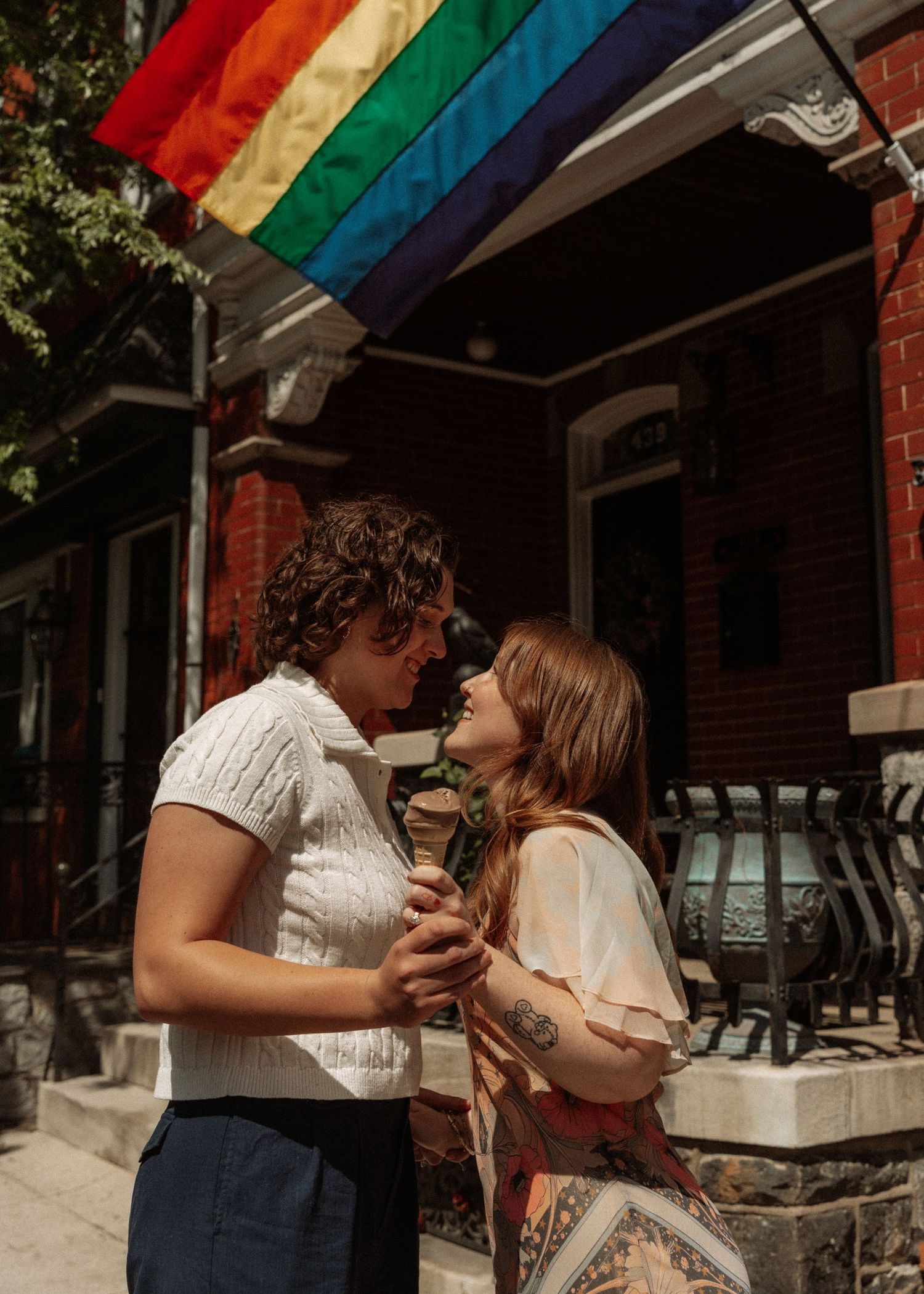 Couple about to kiss under rainbow pride flag outside brick building on sunny day.