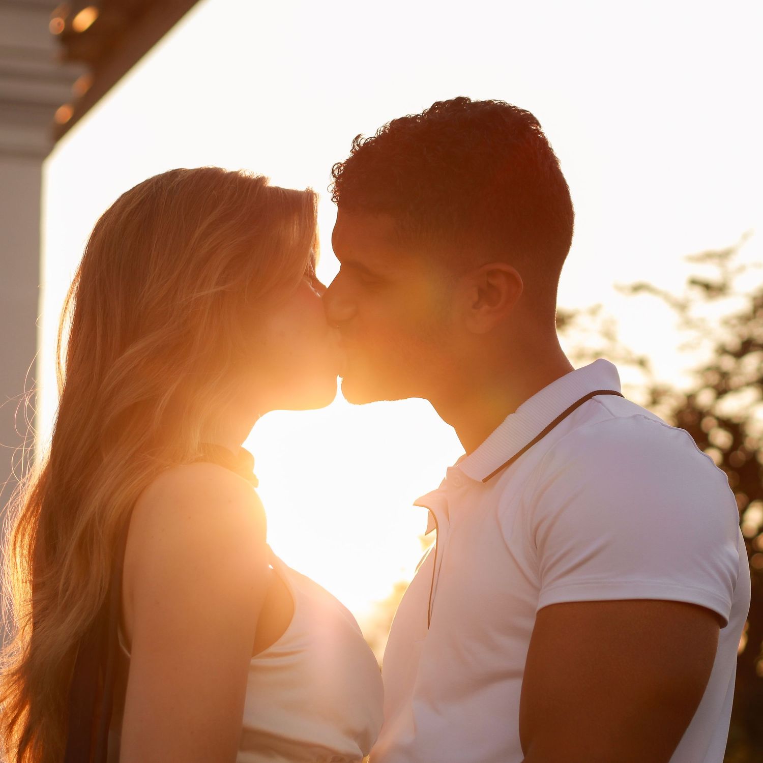 Couple sharing a romantic kiss backlit by golden sunset light outdoors.