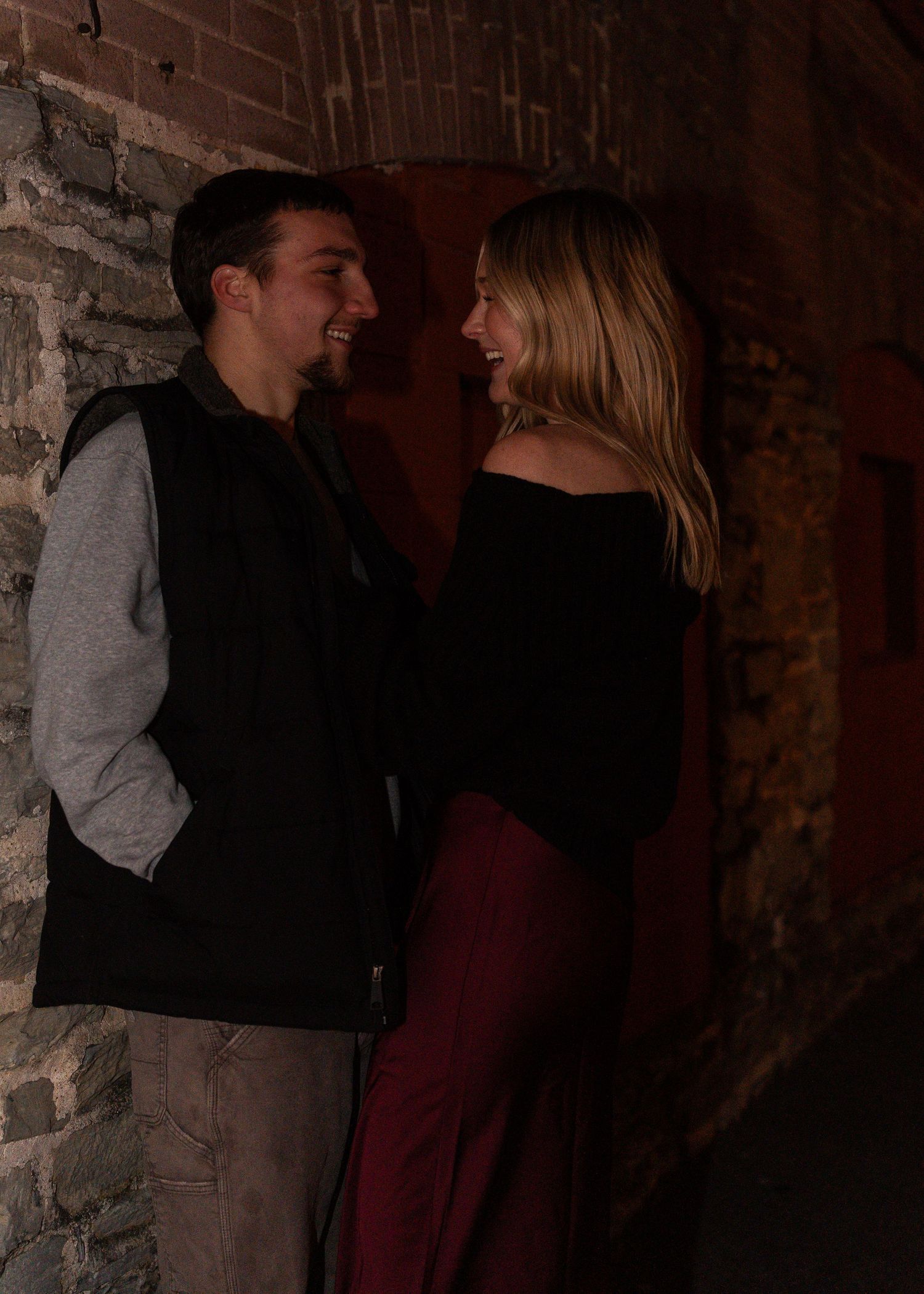 Laughing couple sharing a joyful moment together against a rustic brick wall in warm moody evening light.