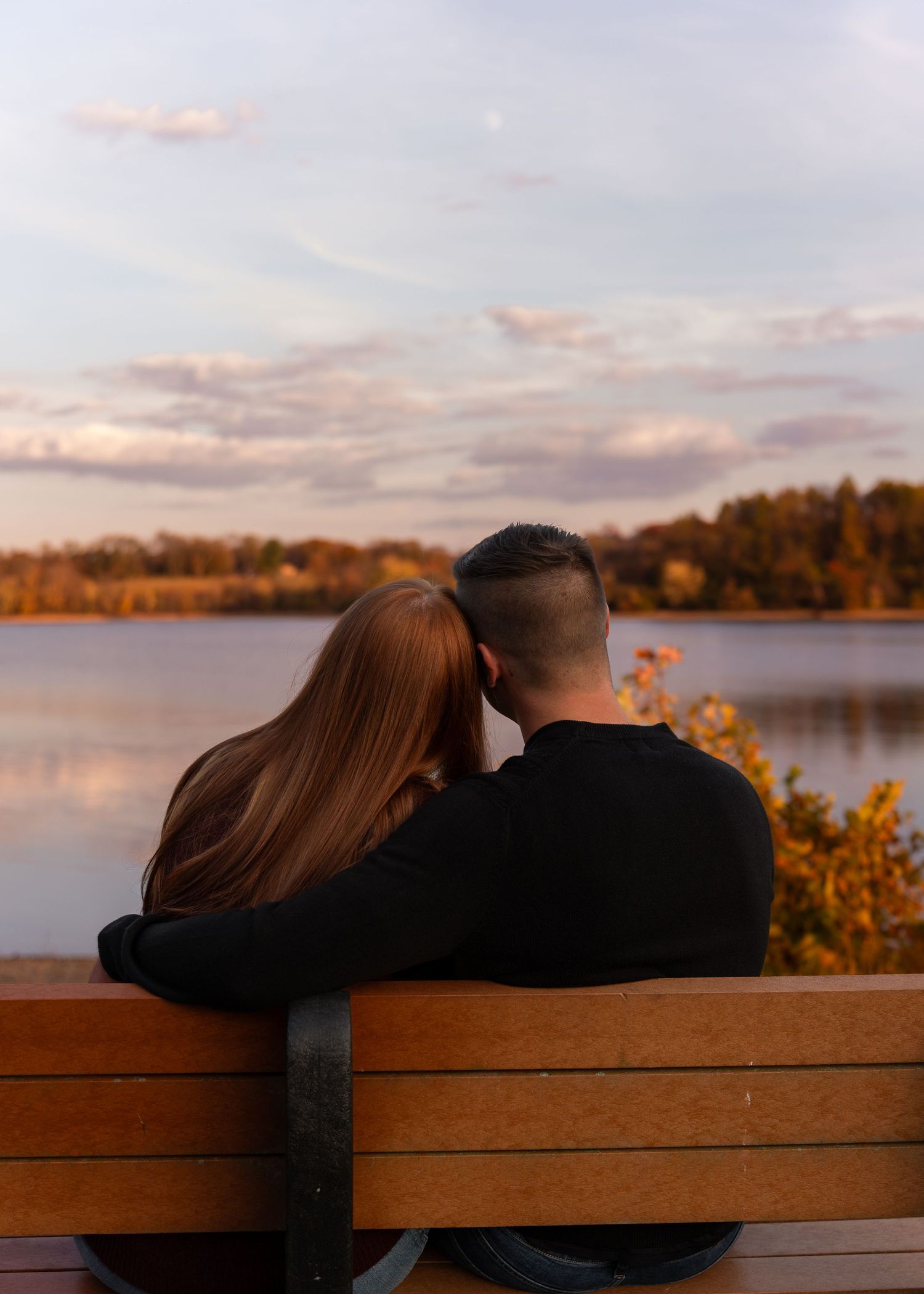 Couple sits on a bench from behind, watching a serene lake reflection under a pastel evening sky.
