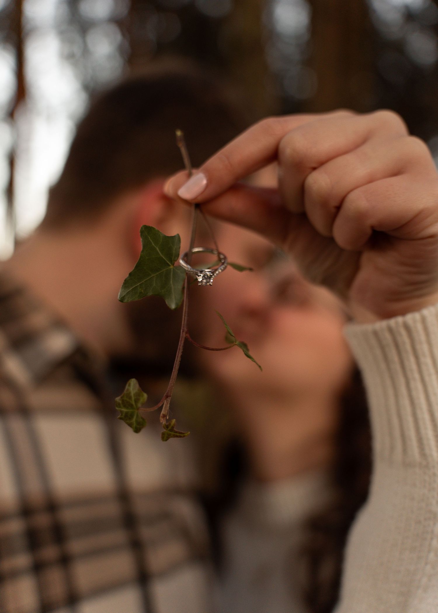 Close-up of engagement ring displayed on an ivy sprig with couple kissing softly in background.