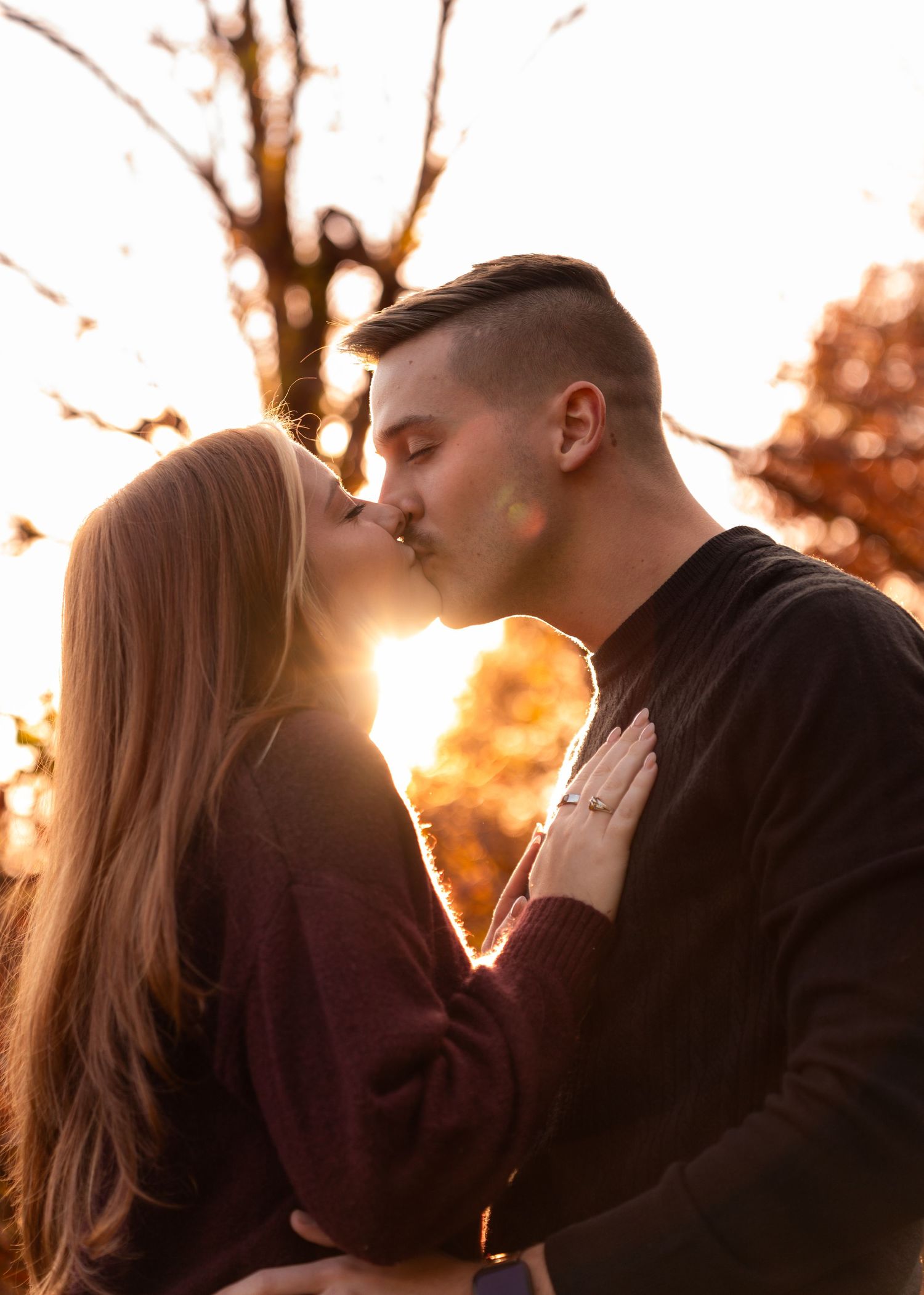 Couple sharing a romantic kiss backlit by a warm golden sunset through autumn trees.