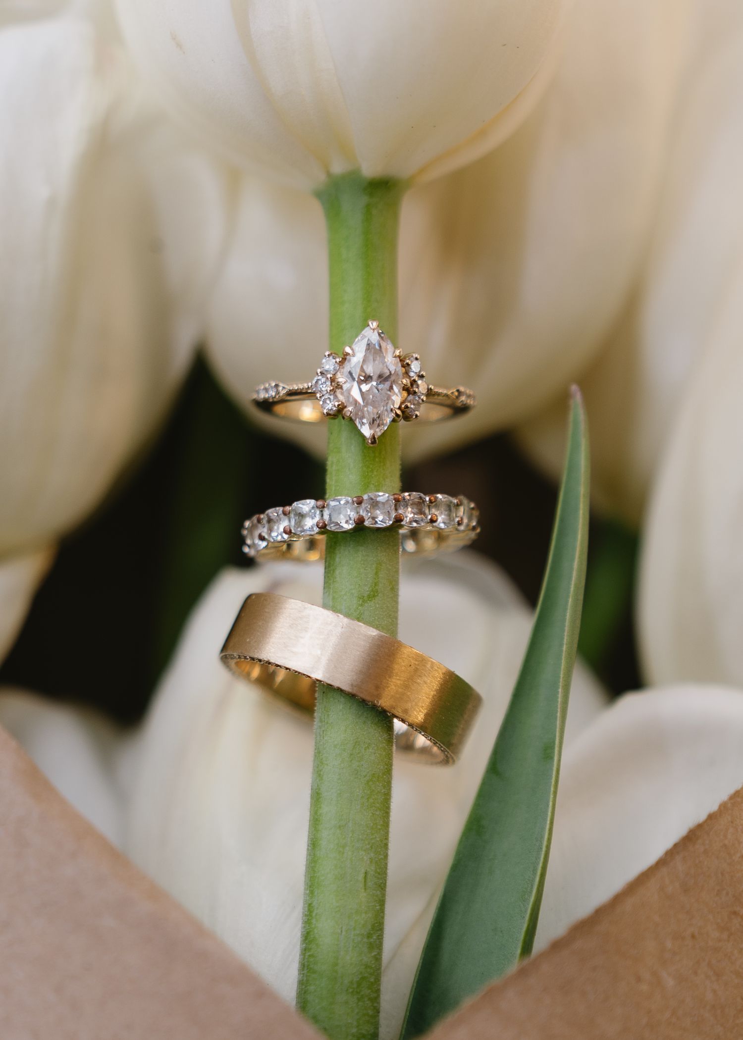 Elegant marquise diamond engagement ring with diamond band and gold wedding band resting on white tulip stems.