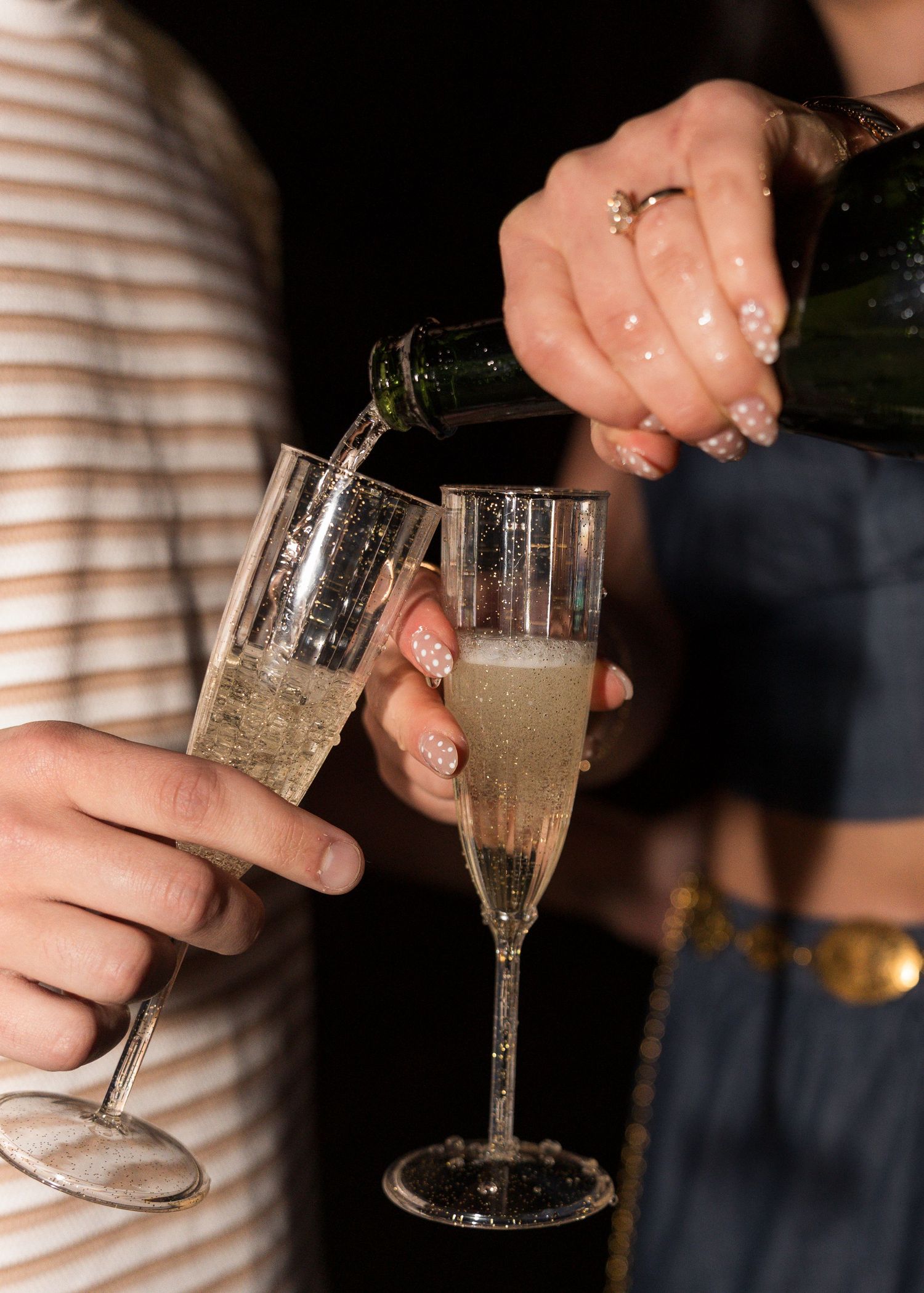 Couple pouring champagne into flutes at a celebration, woman with glitter nails and engagement ring.