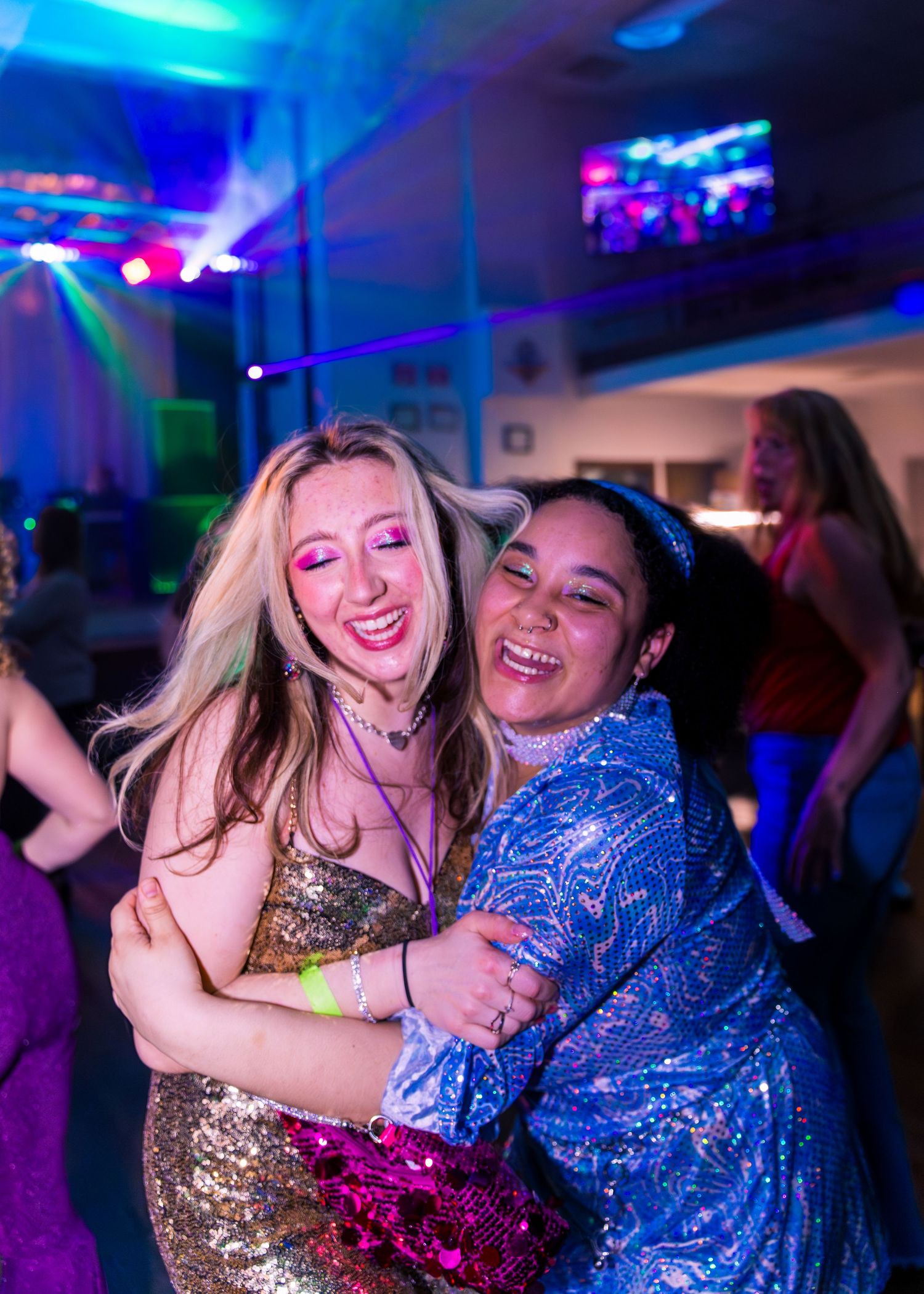 Two women in sequin dresses hugging and laughing at a colorful disco party.