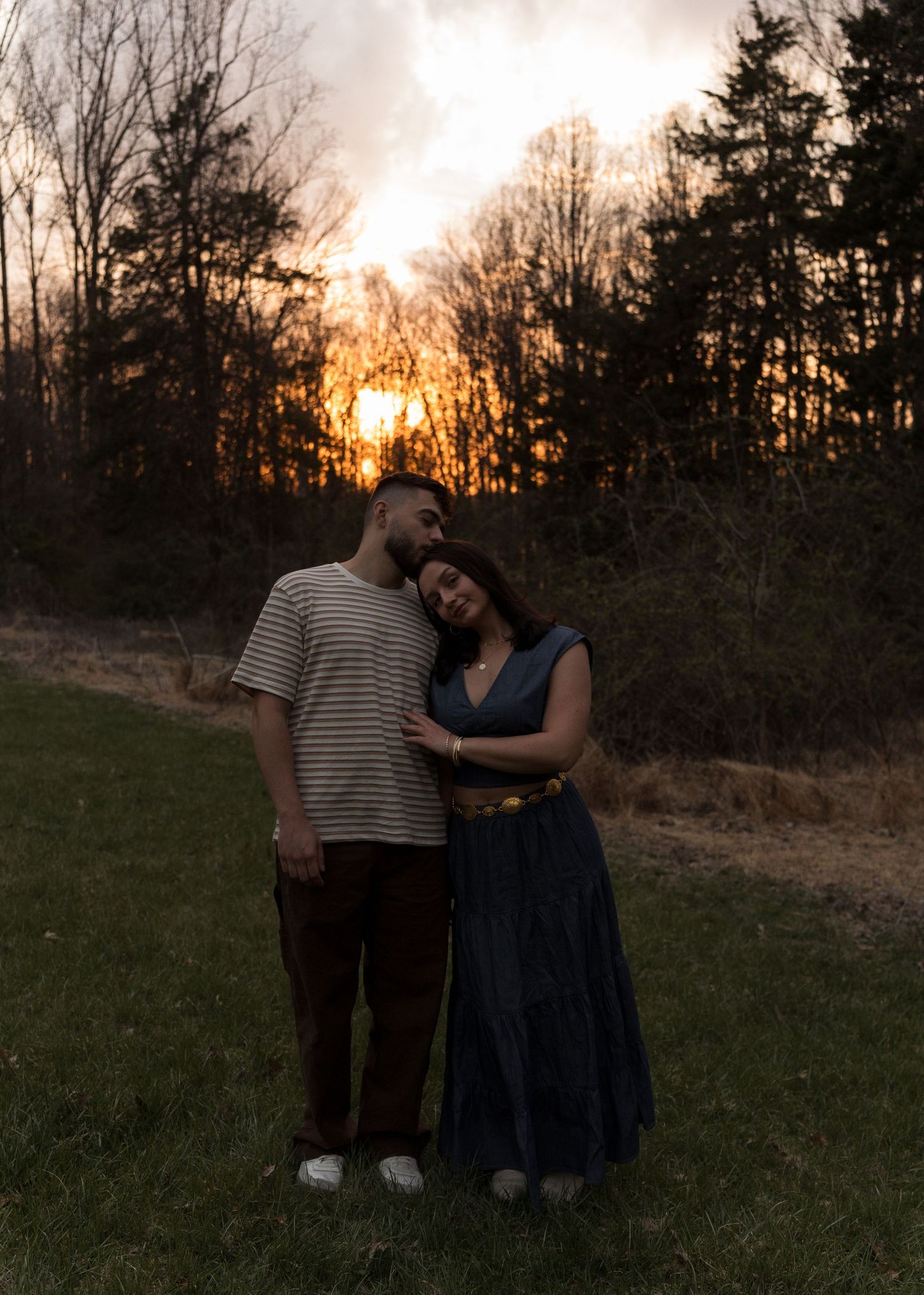 Couple embracing at golden sunset in field, woman in navy dress leaning on partners shoulder.