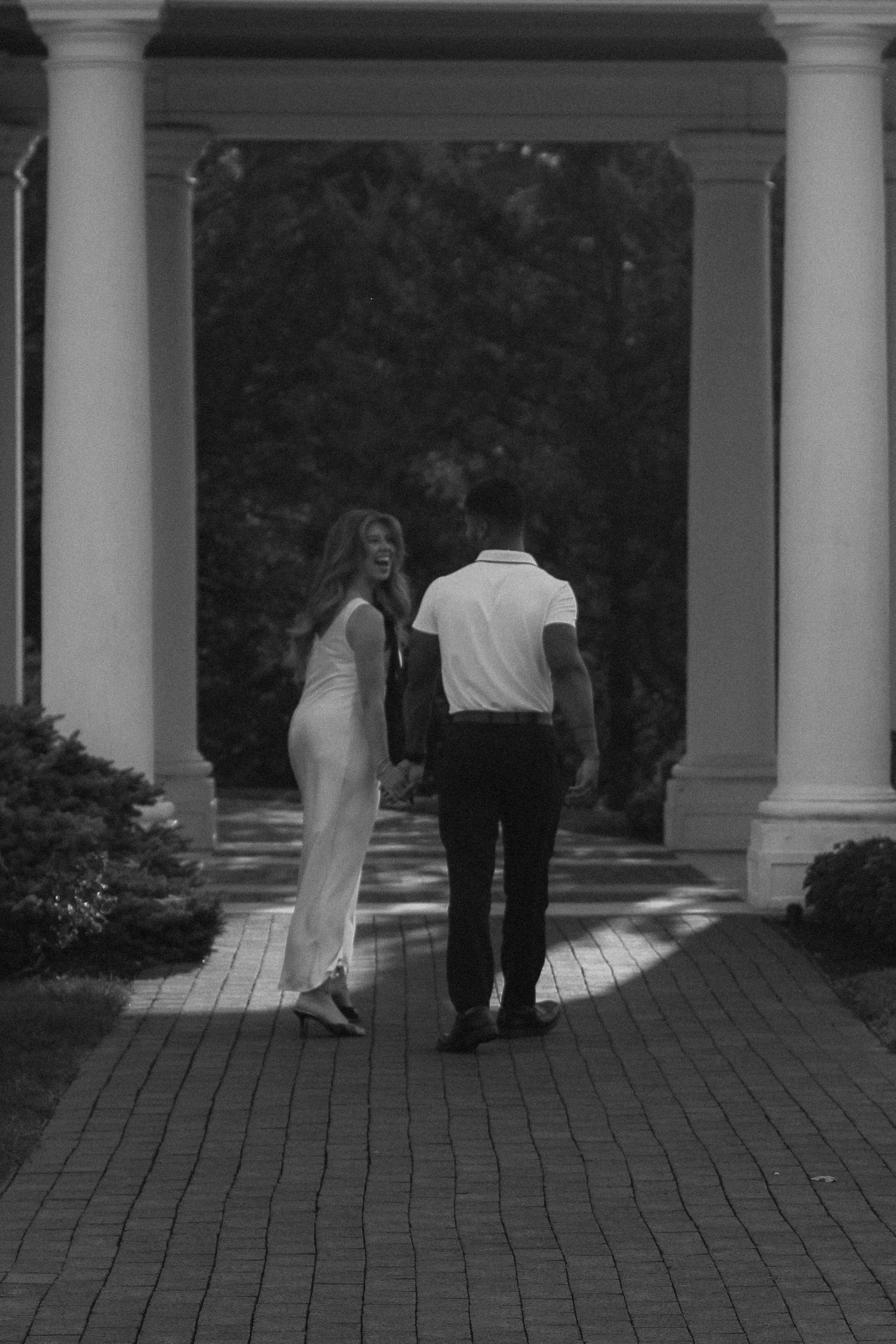 Black and white photo of couple holding hands walking through elegant columned pathway.