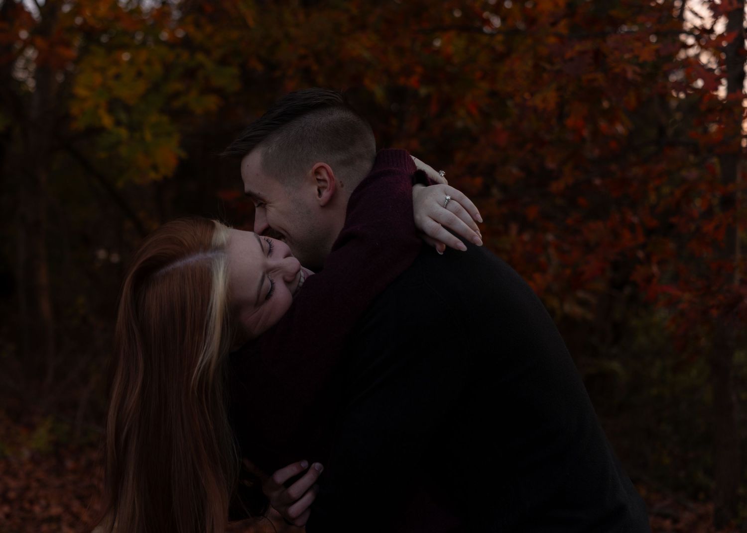 Romantic couple embracing and kissing outdoors surrounded by vibrant autumn foliage at dusk.