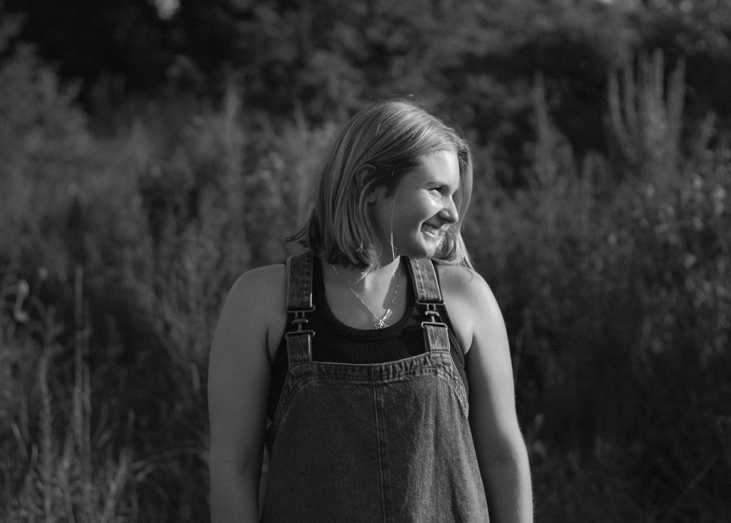 Black and white portrait of a smiling young woman in overalls standing in a natural outdoor setting.