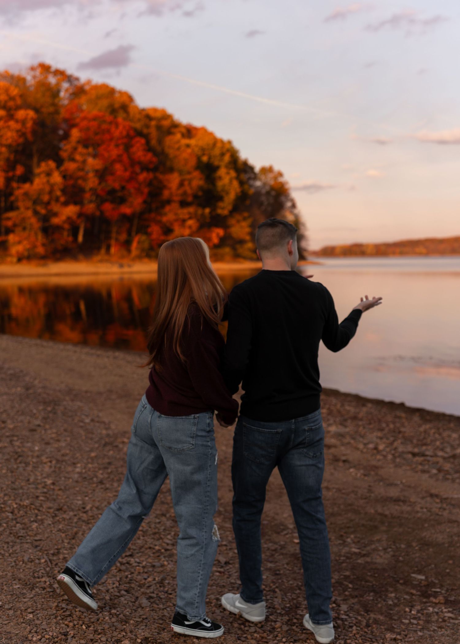 Couple walking hand-in-hand along a lakeside shore surrounded by vibrant fall foliage at sunset.