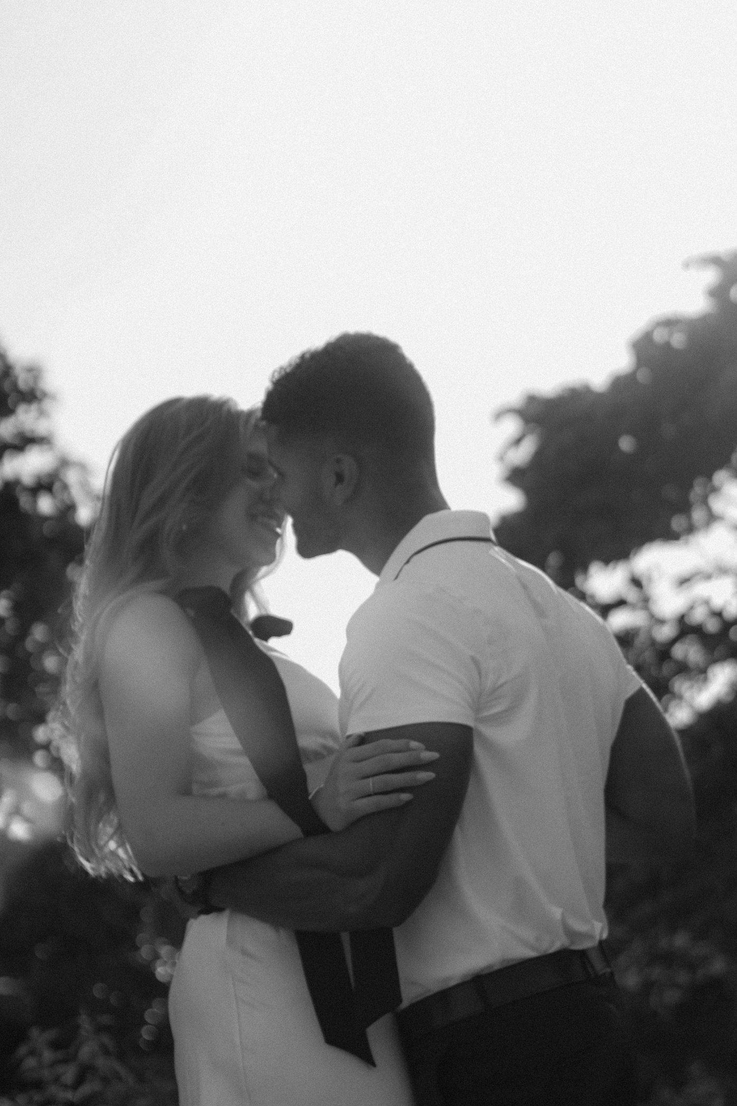 Black and white photo of an interracial couple embracing and almost kissing outdoors among trees.