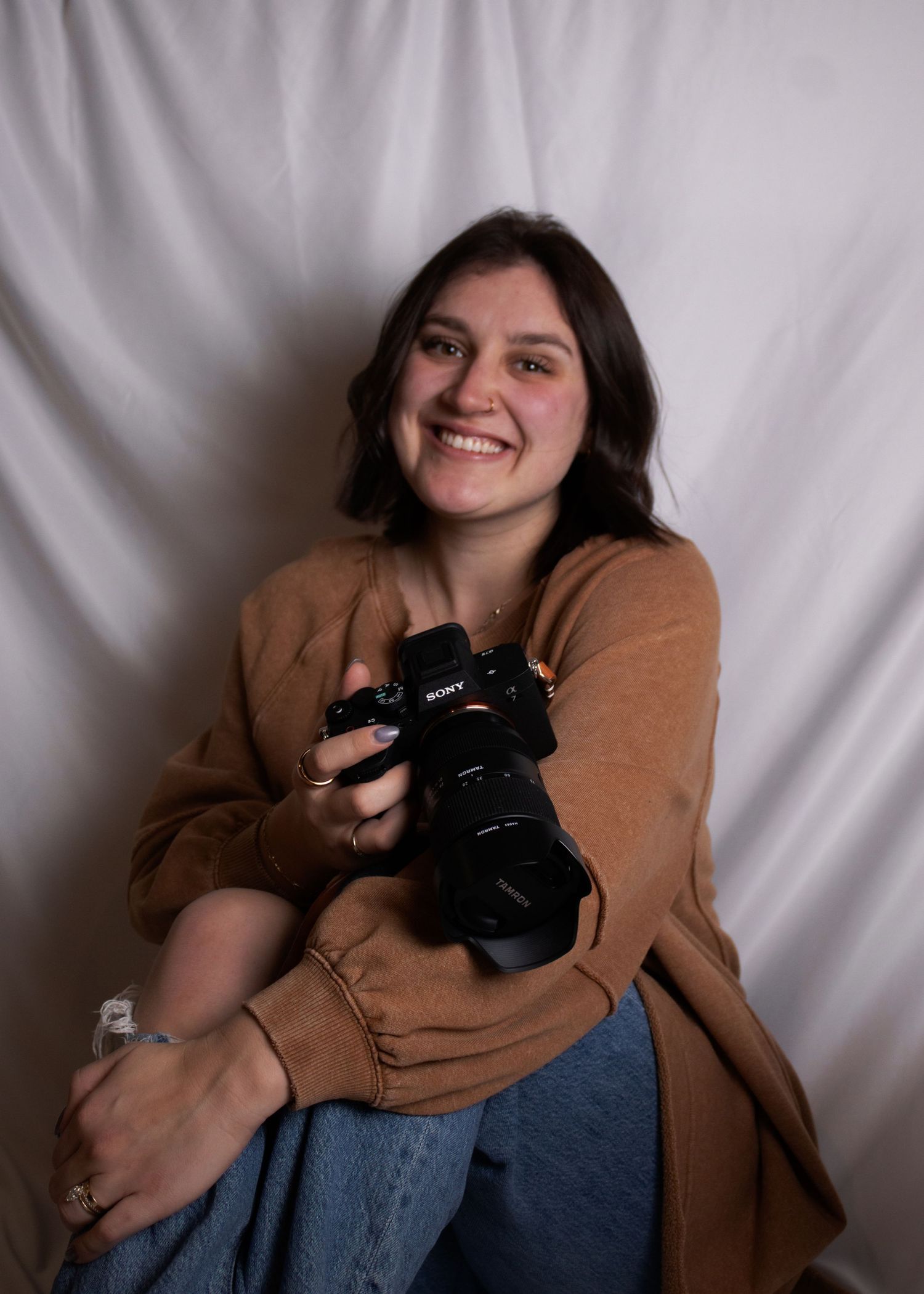 Smiling young female photographer sitting with Sony camera against white fabric backdrop.