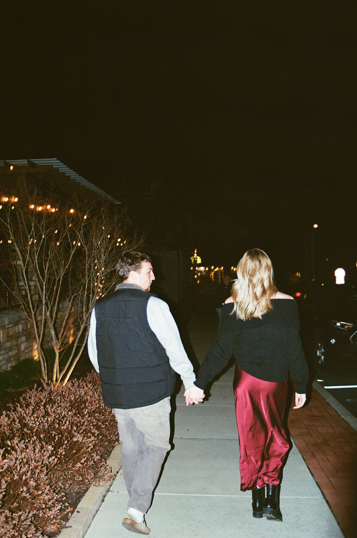 Couple holding hands walking along a nighttime sidewalk, woman in red satin skirt and black top.