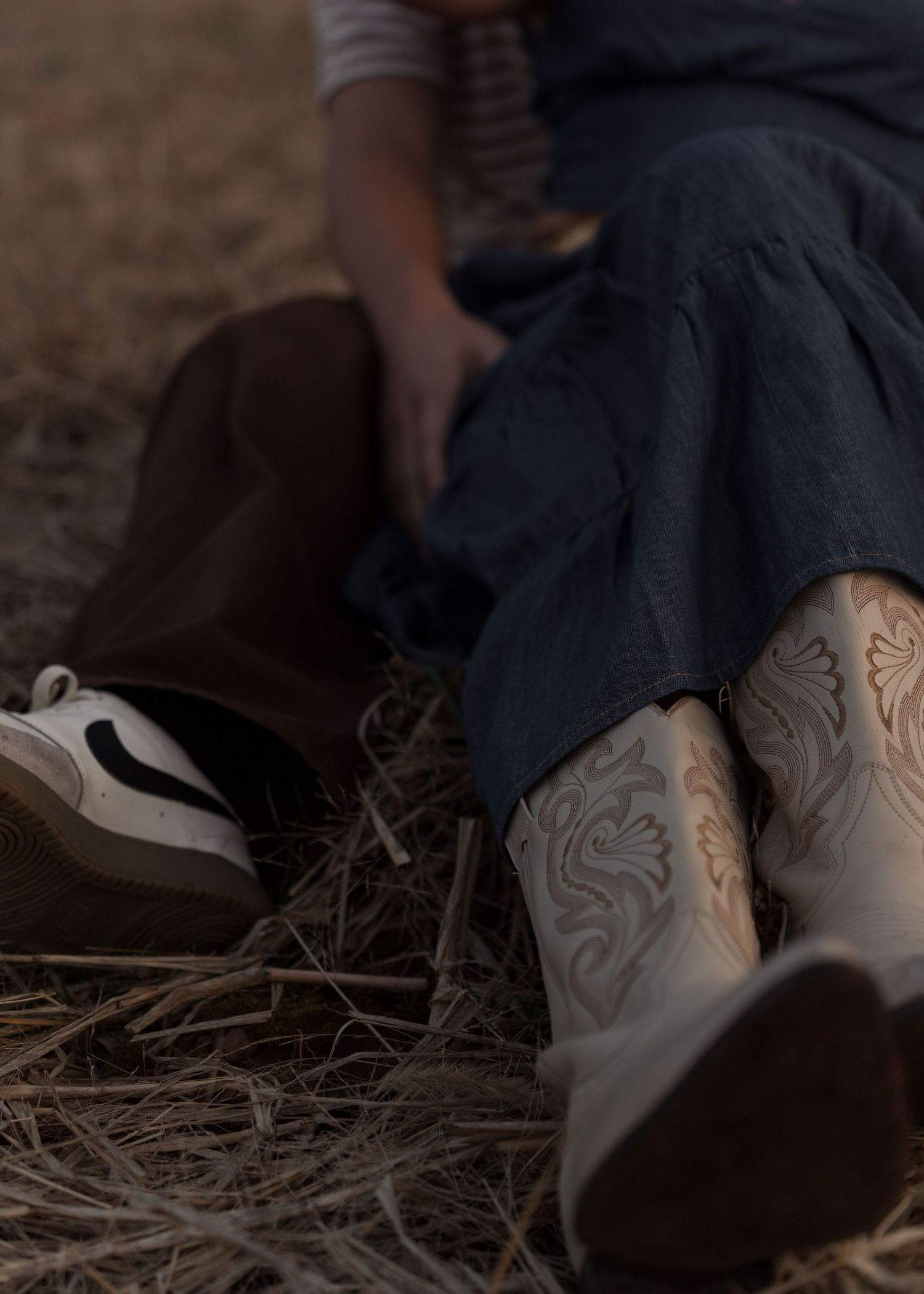 Close-up of couples feet, woman in embroidered cowboy boots and navy skirt sitting in hay.