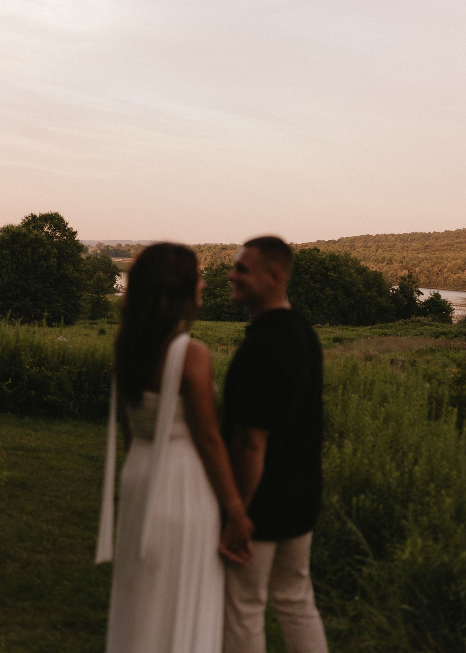 Couple holding hands in a lush green field at sunset, woman in white dress and man in black shirt.