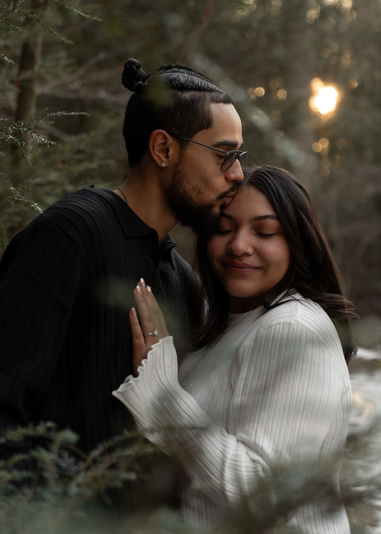 Man kissing woman on temple in forest at golden hour, couple embracing romantically among pine trees.