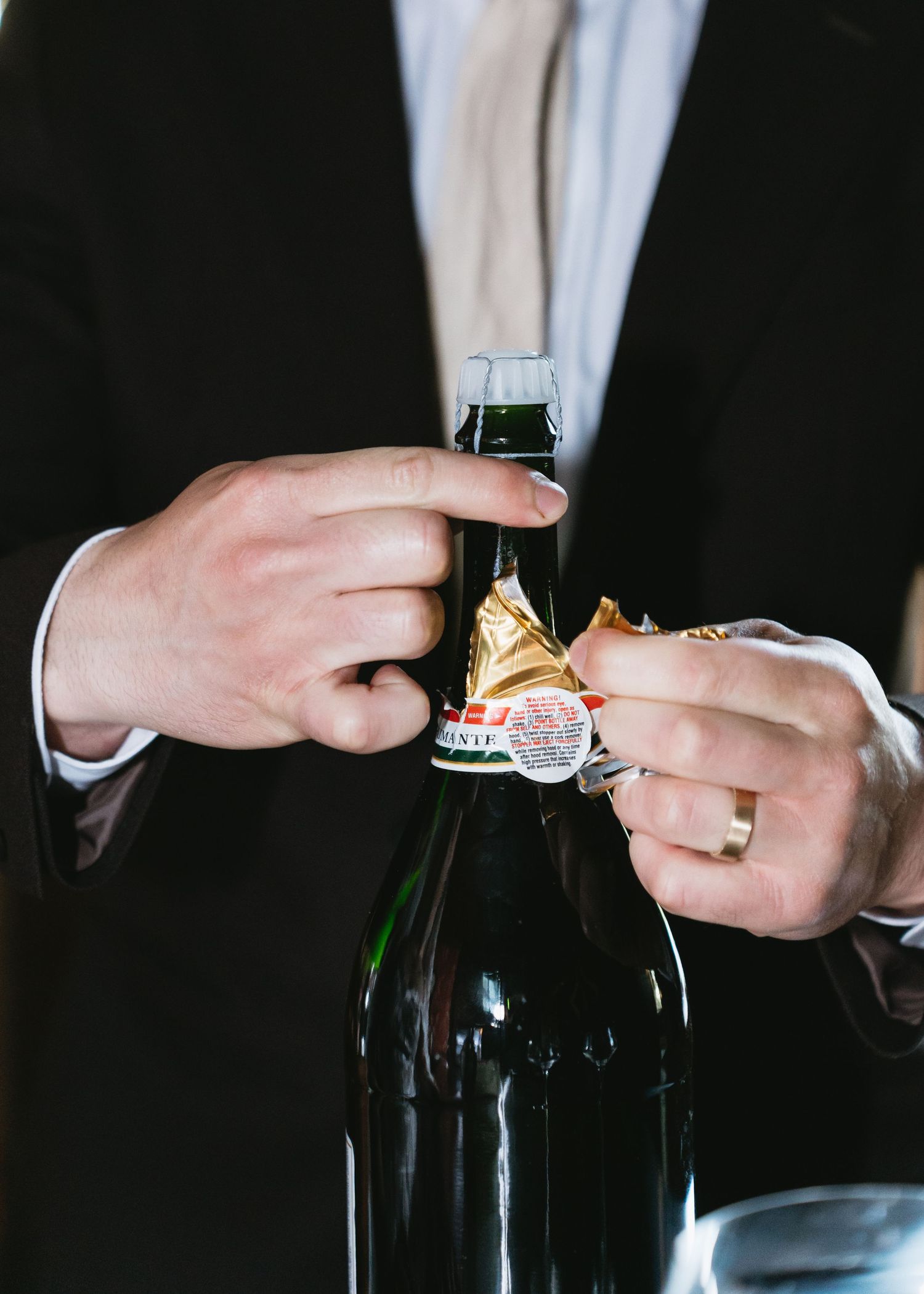 Groom in black suit opening a bottle of Perrier-Jouet champagne with wedding ring visible on his hand.
