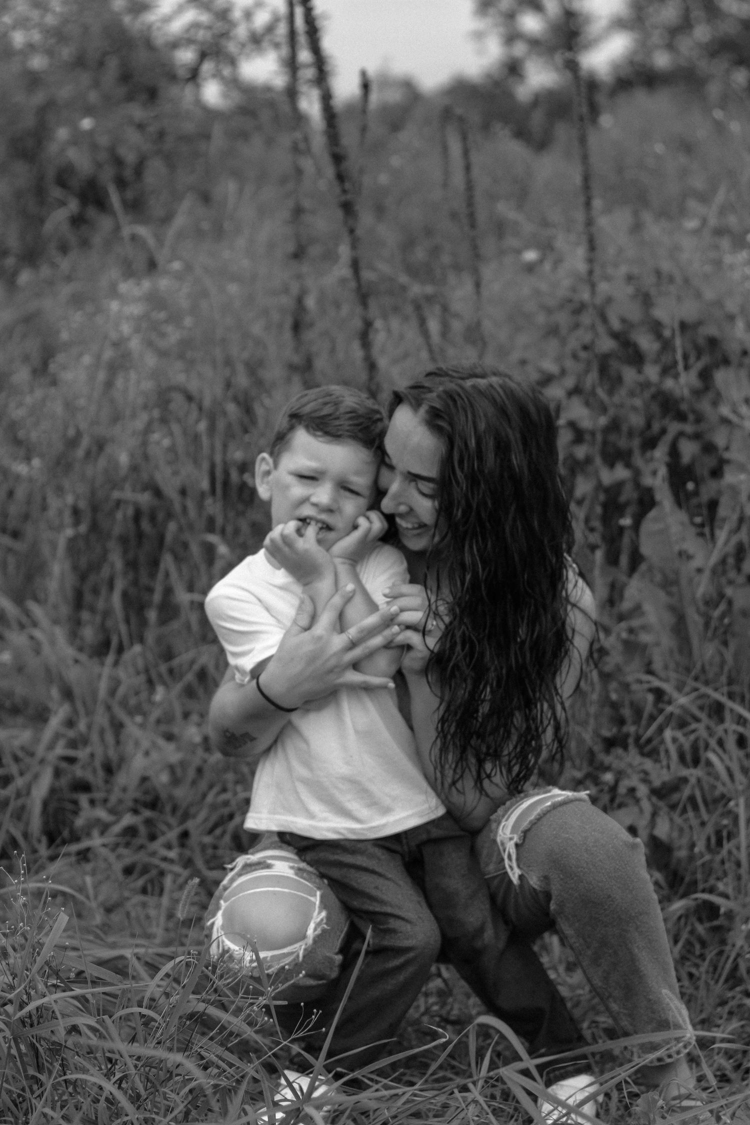Black and white photo of a mother hugging her young son in a lush outdoor field setting.