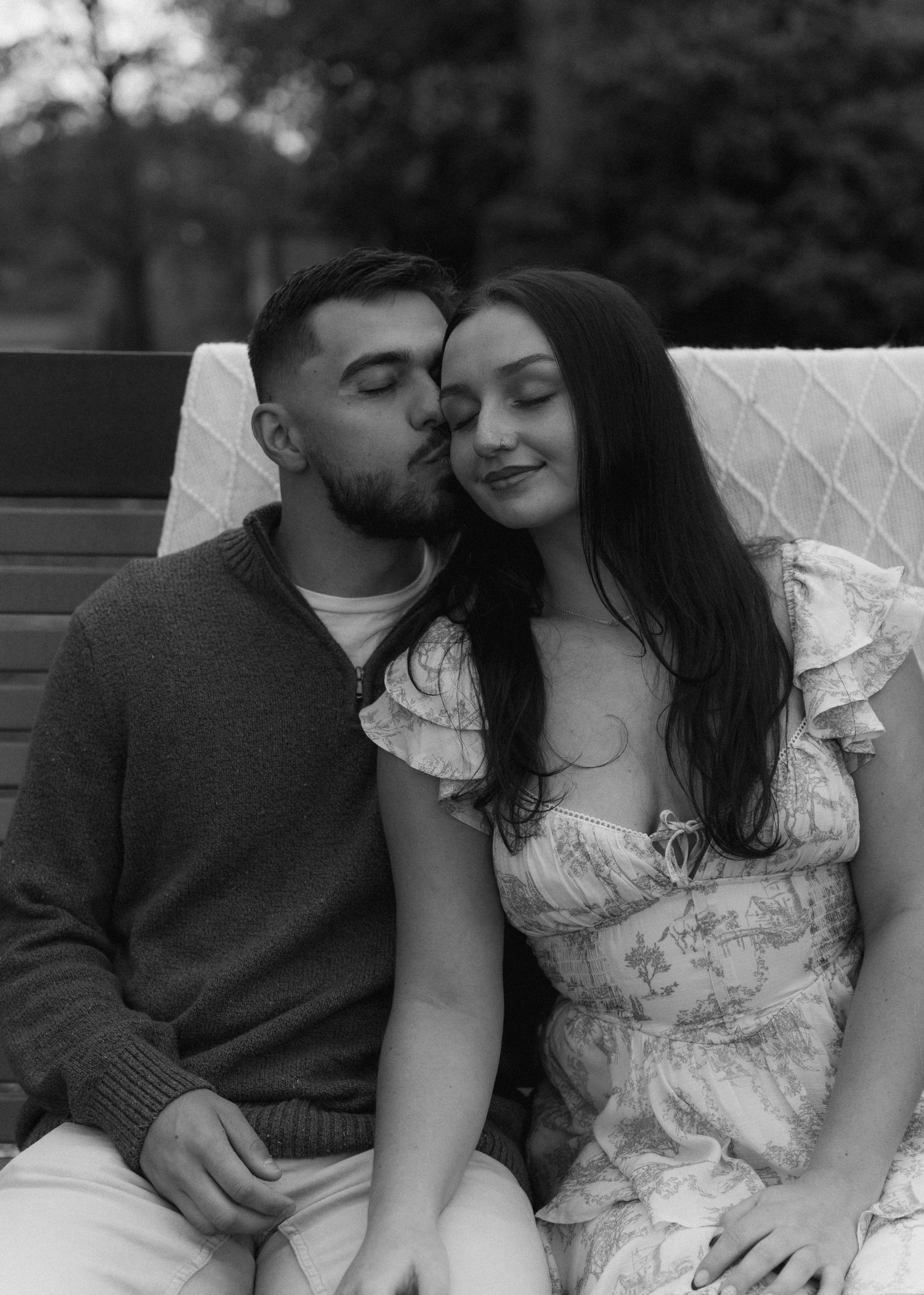 Black and white photo of a couple sitting on a bench as man kisses womans cheek.