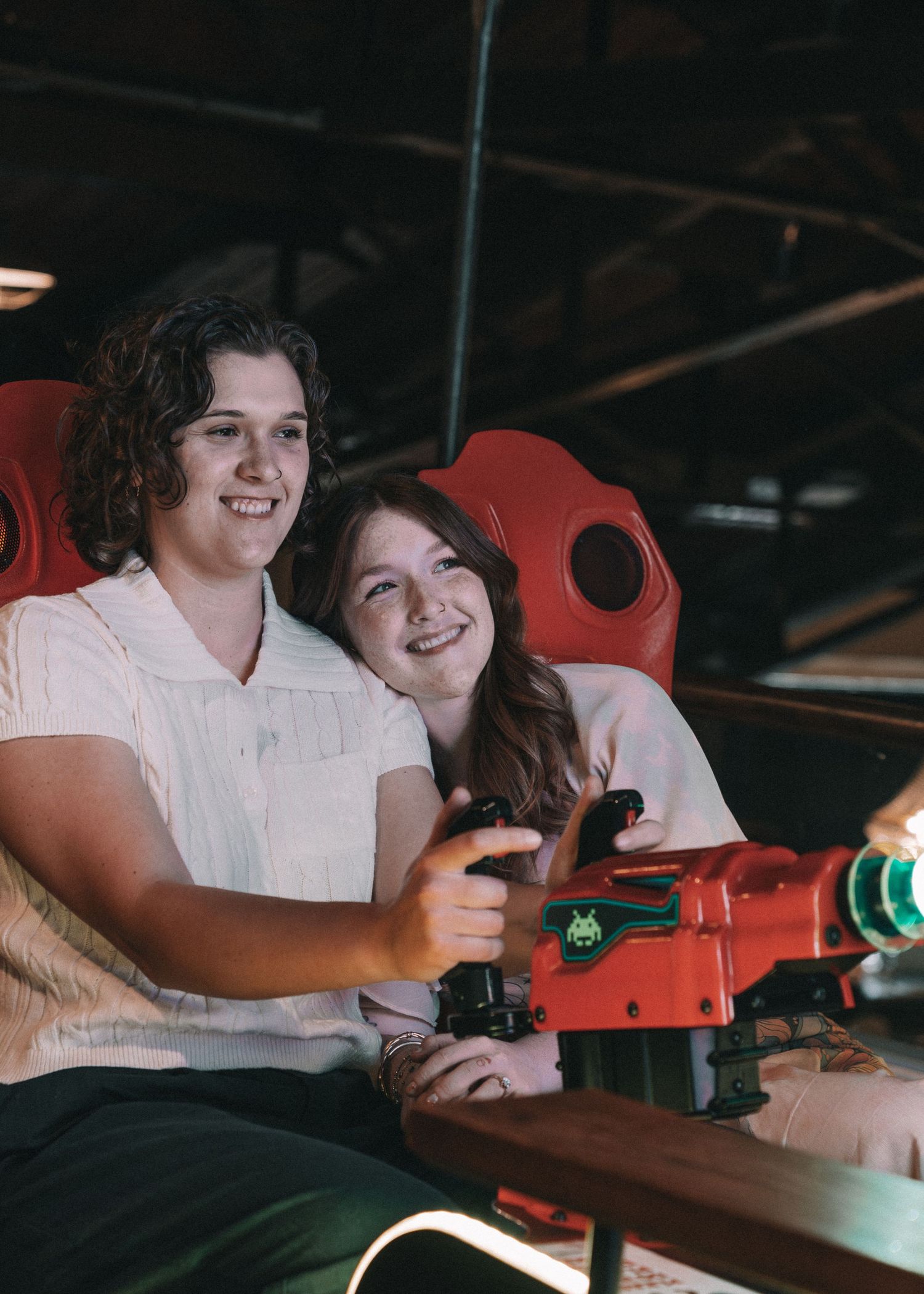 A smiling couple enjoys an arcade shooting game together, seated in red chairs in a dimly lit venue.
