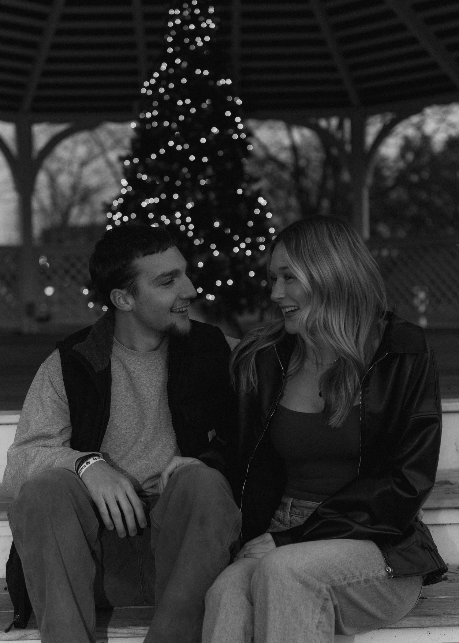 Couple sitting together laughing in black and white photo with Christmas tree and gazebo behind them.