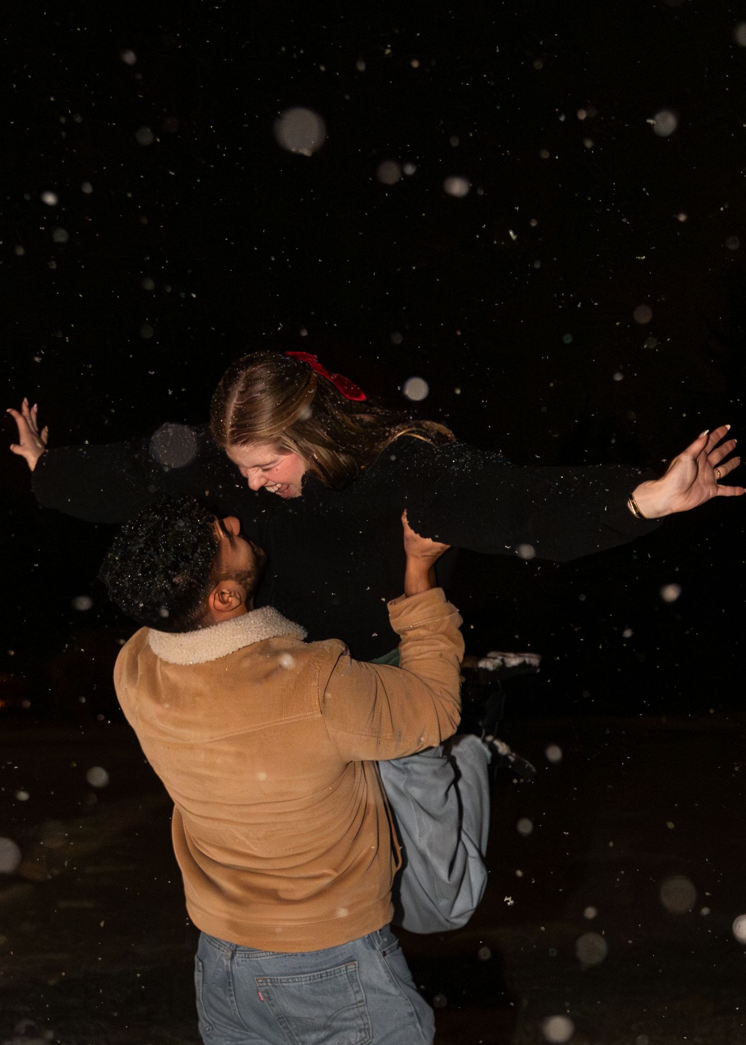 Man joyfully lifting woman with outstretched arms in snowy night, both laughing in winter snowfall.