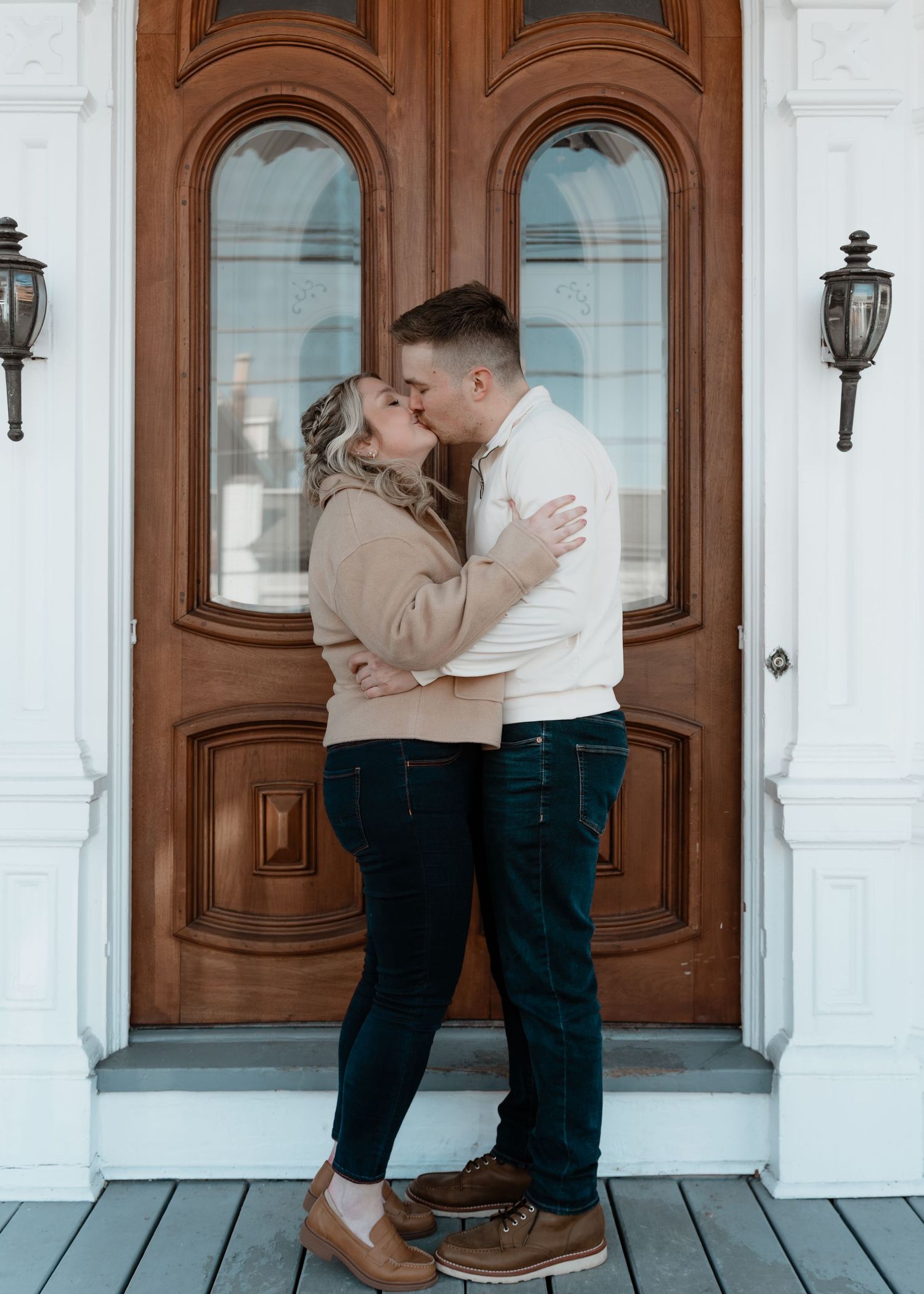 Couple sharing a romantic kiss in front of an elegant wooden arched door on a charming porch.