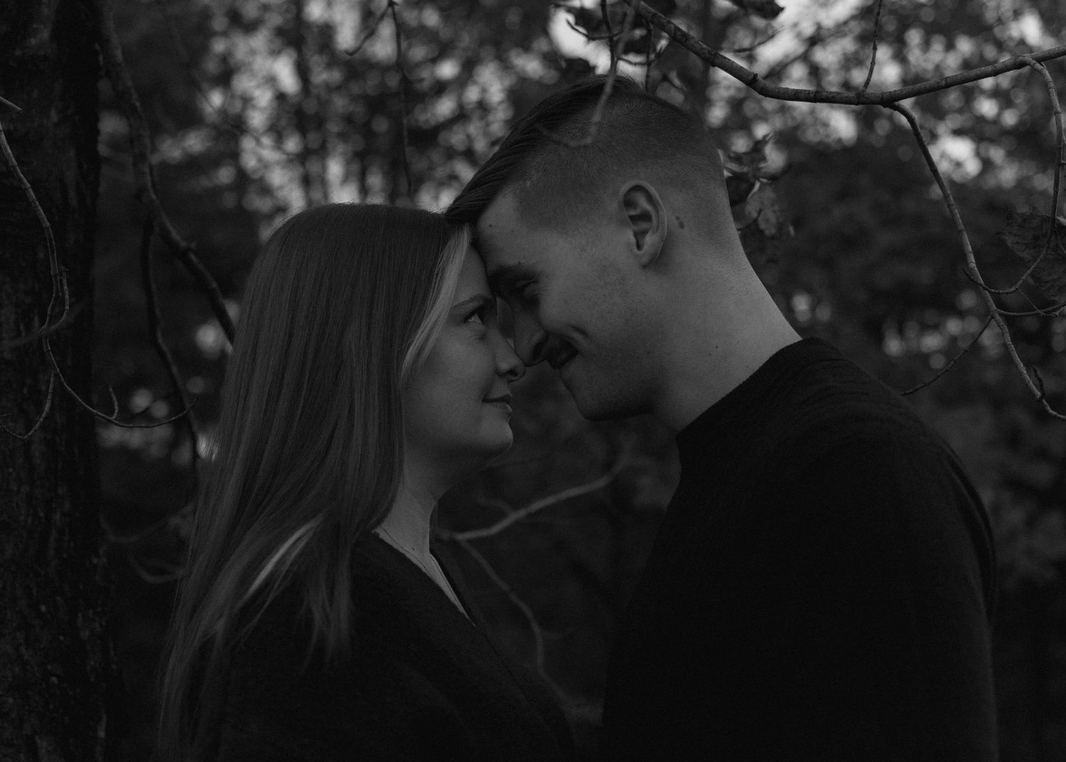 Black and white photo of couple facing each other intimately under tree branches in forest.