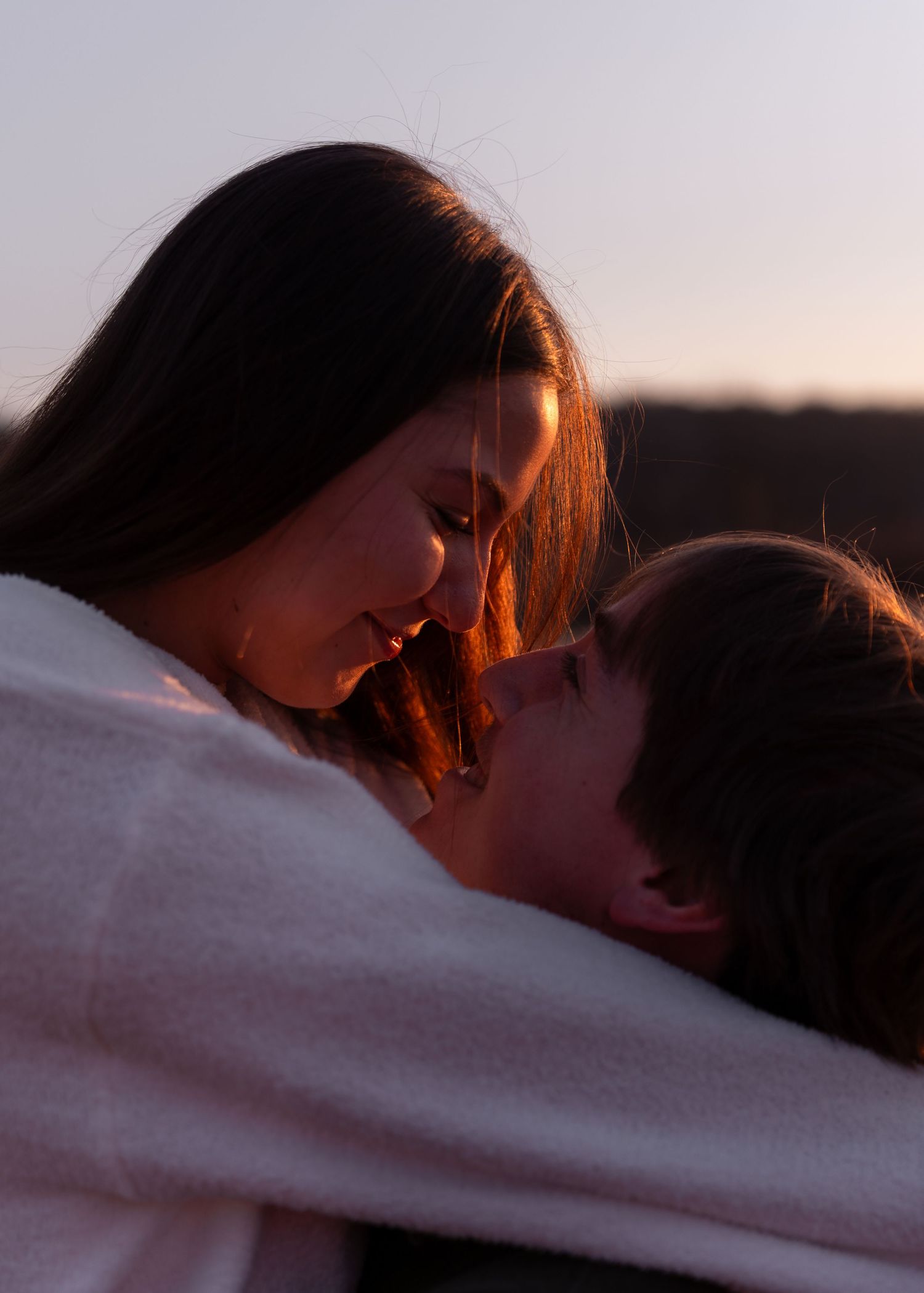 Couple wrapped in a white blanket sharing a tender moment at sunset in an open field.